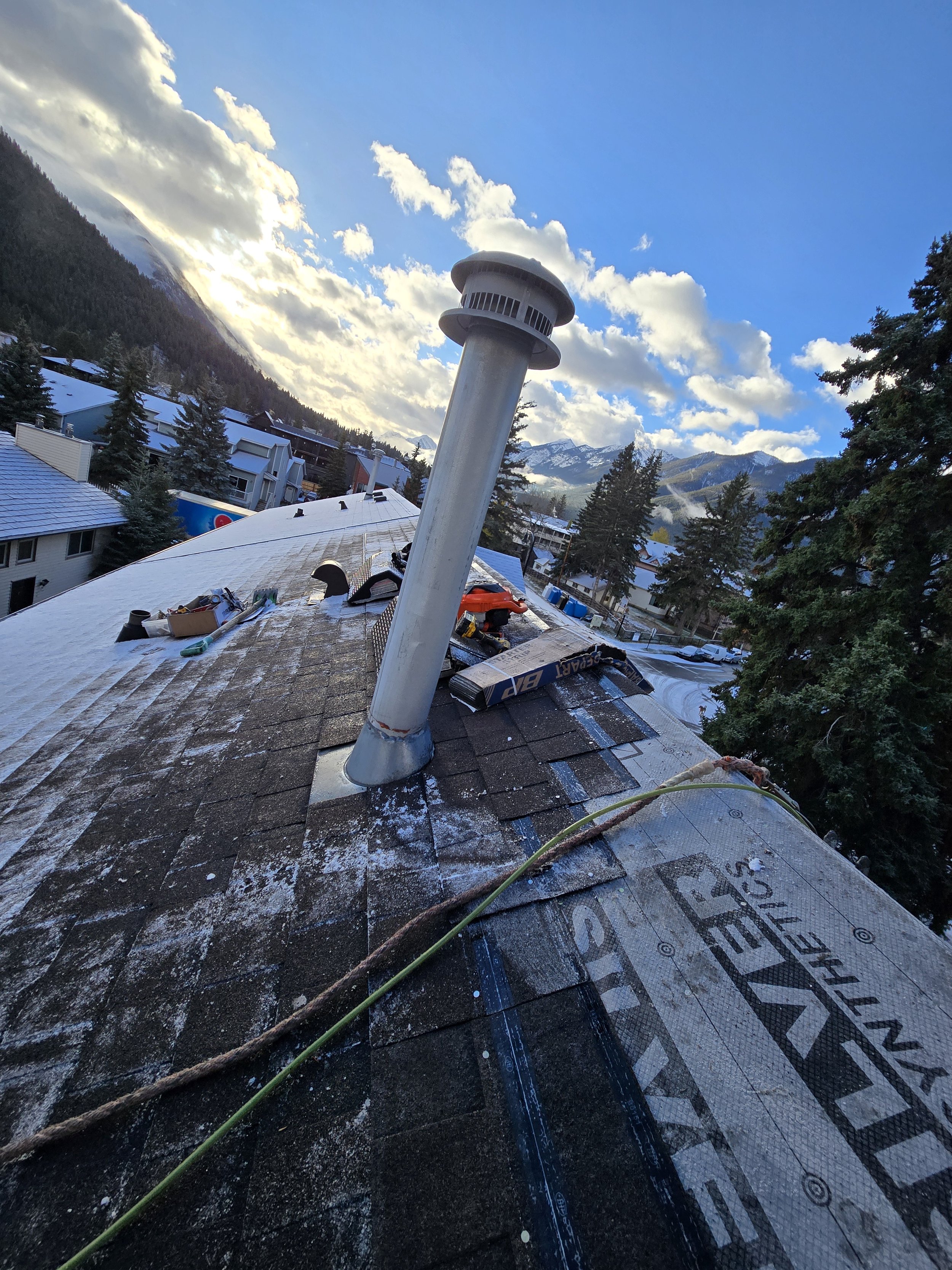 A rooftop with construction tools and materials, including a large vent pipe, tools, and insulation, against a backdrop of a mountain town with snow-capped peaks and a partly cloudy sky.