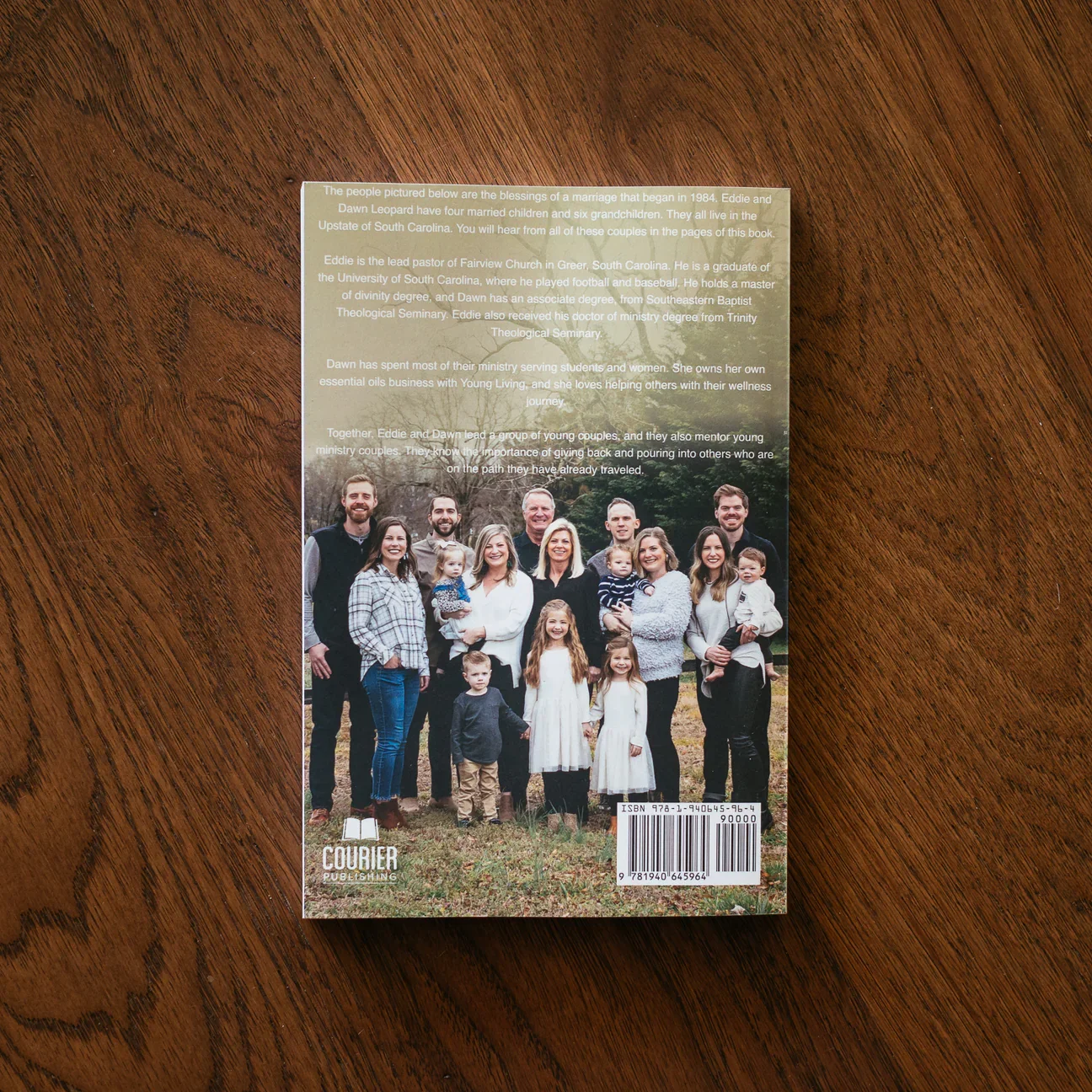 Back cover of a family photo book with a group photo of multiple family members and children in an outdoor setting, along with a description of the family members and their background, and a barcode at the bottom right corner.
