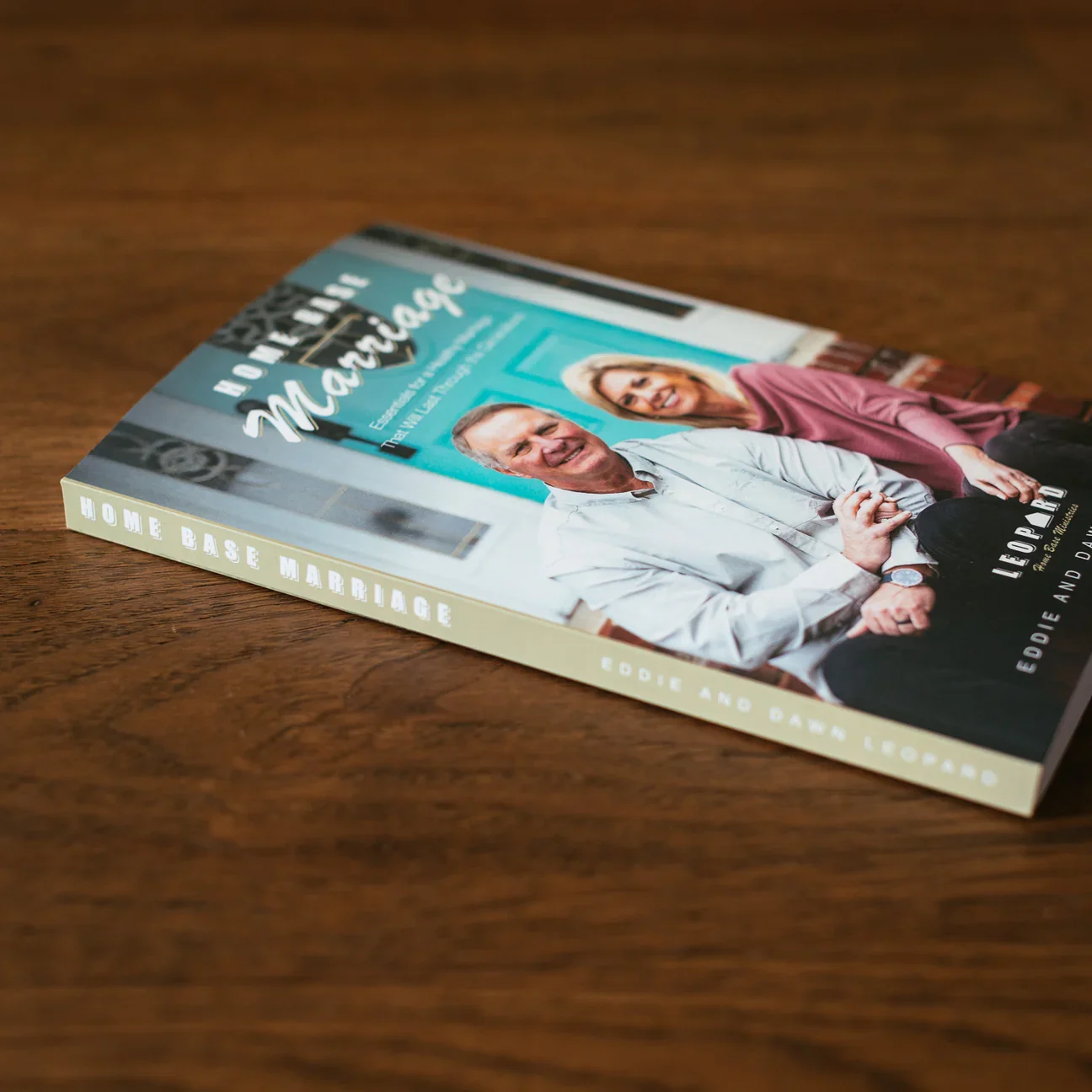A hardcover book titled "Home Base Marriage" by Eddie and Banne Leonard lies on a wooden surface. The cover features a photo of a smiling middle-aged couple sitting together in front of a teal-colored door.