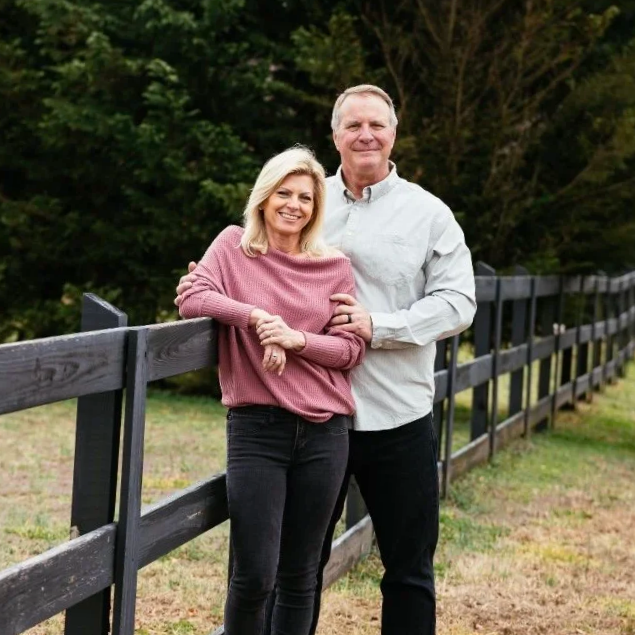 A smiling woman with blonde hair wearing a pink sweater and black pants standing next to a smiling man with gray hair wearing a white shirt and black pants, outdoors by a black fence with trees in the background.