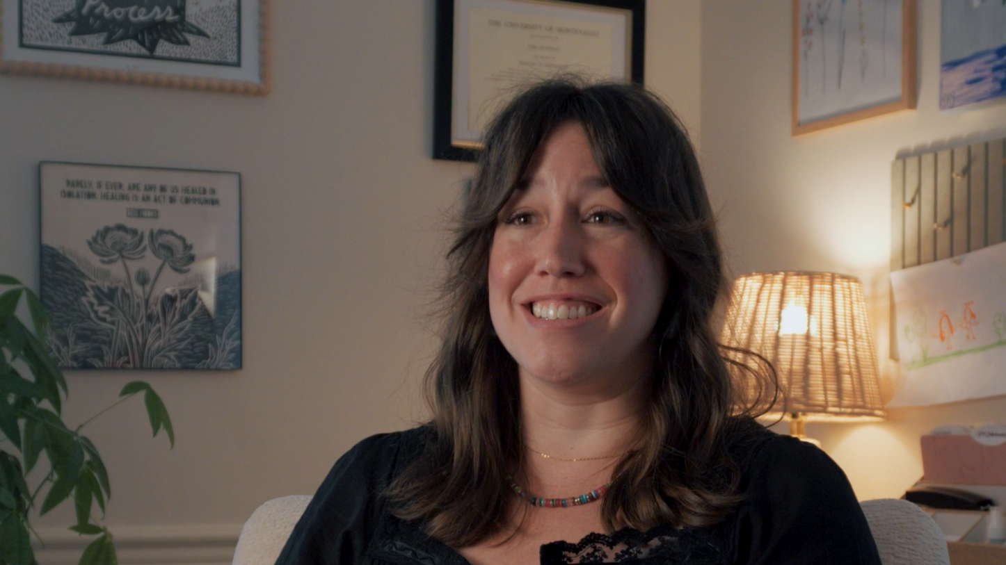 A woman with shoulder-length dark hair smiling in a cozy room with framed artwork and a lamp behind her.