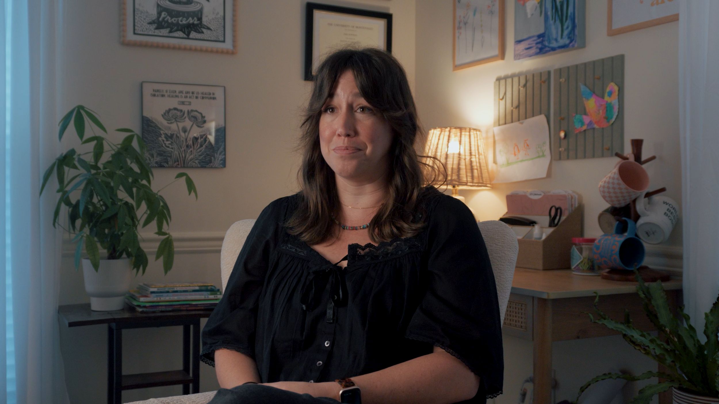 A woman with dark hair and a colorful beaded necklace sitting in a cozy, art-filled room, with a bookshelf, plants, and colorful artwork behind her.