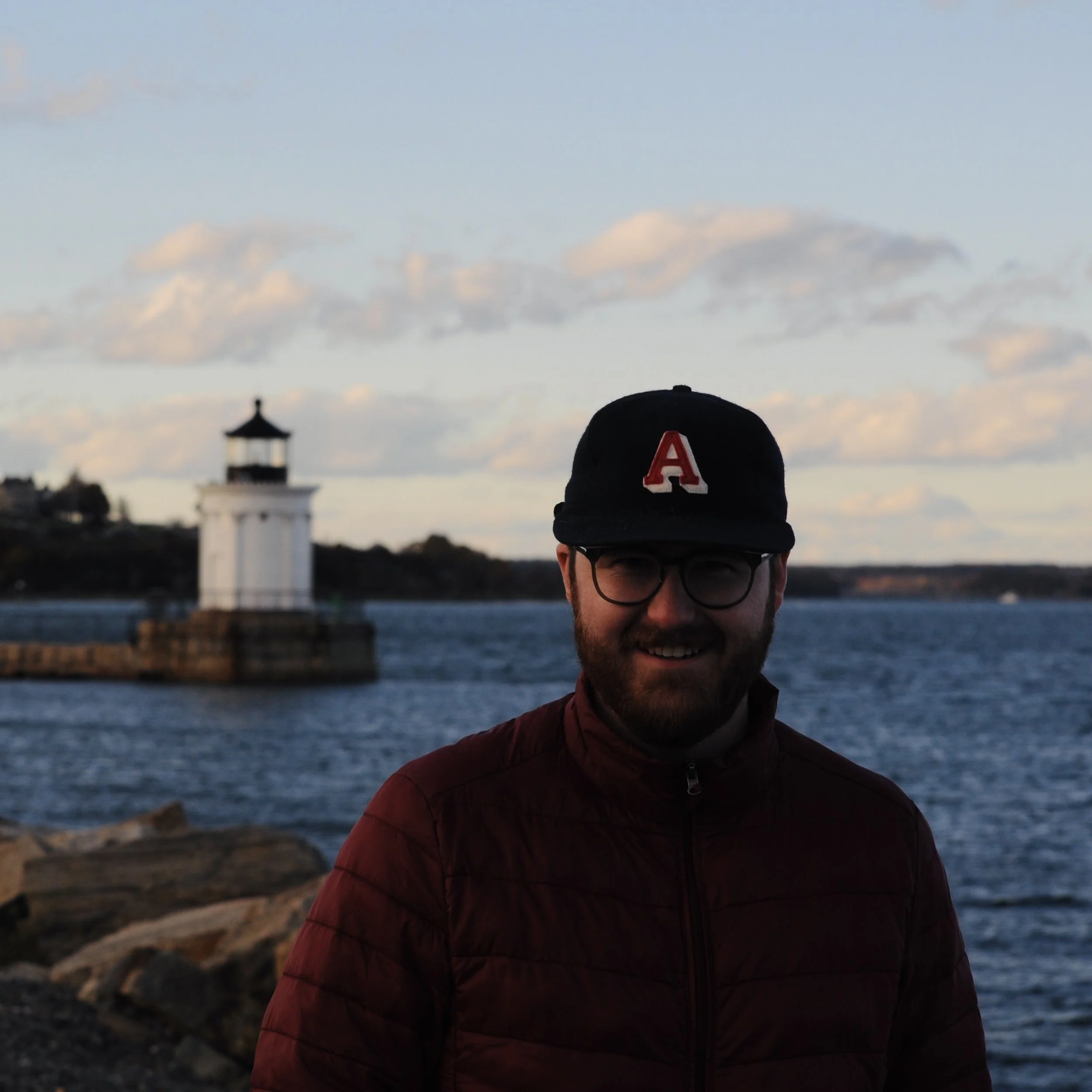 A man with glasses and a beard wearing a black cap with a red letter A and an orange jacket is standing outdoors near water, with a lighthouse and cloudy sky in the background during evening or early night.