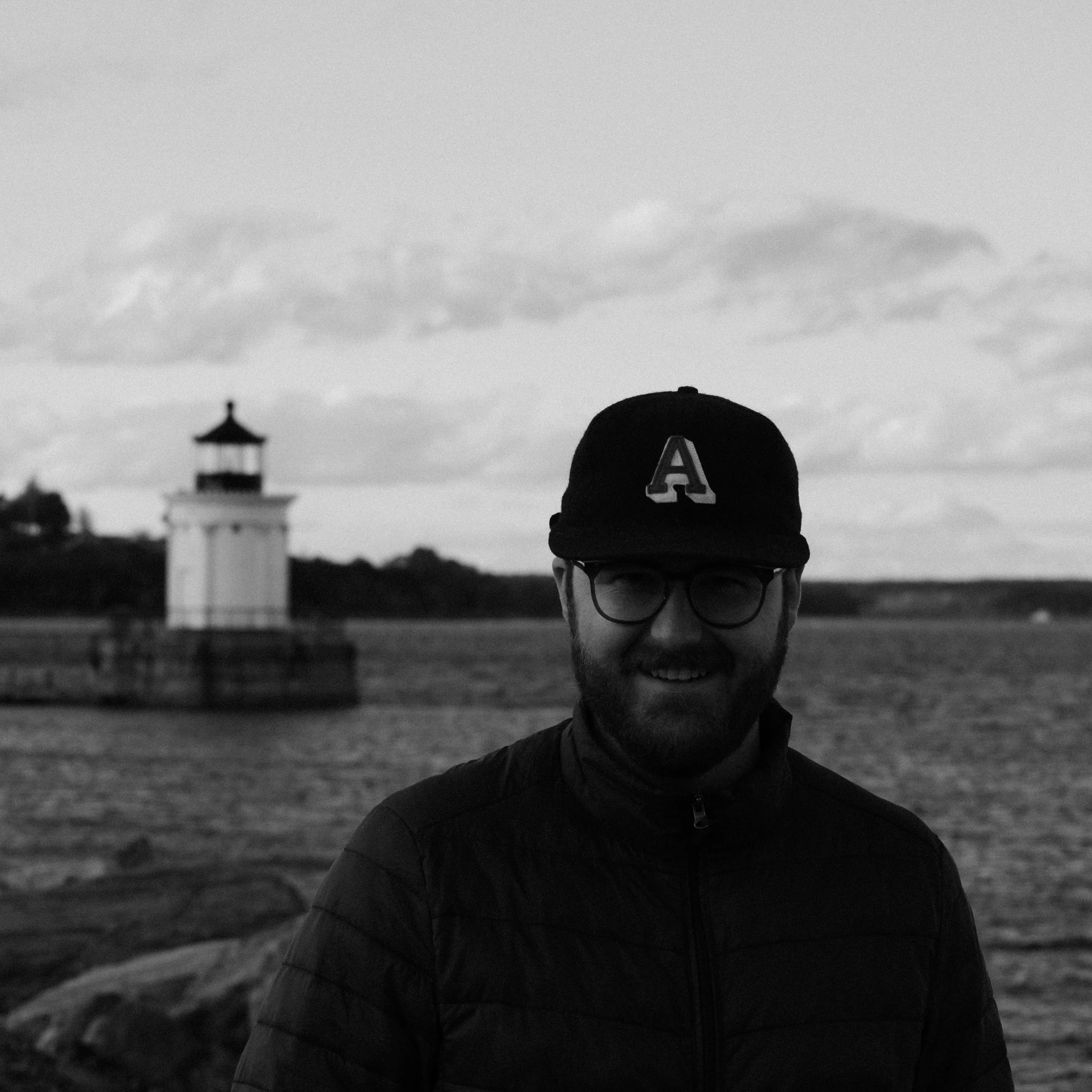 A person wearing glasses, a baseball cap with an 'A' logo, and a jacket, standing outdoors with cloudy sky and a lighthouse in the background.