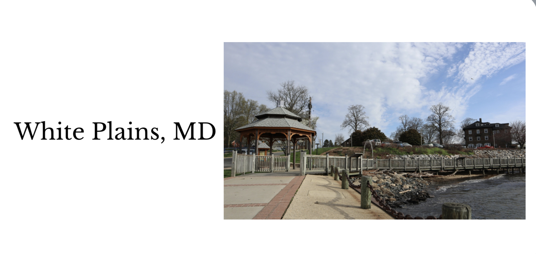 A seaside promenade in White Plains, Maryland, featuring a wooden gazebo, a paved walkway, a rock-stone shoreline, and a parking area with cars, under a partly cloudy sky with trees and a building in the background.
