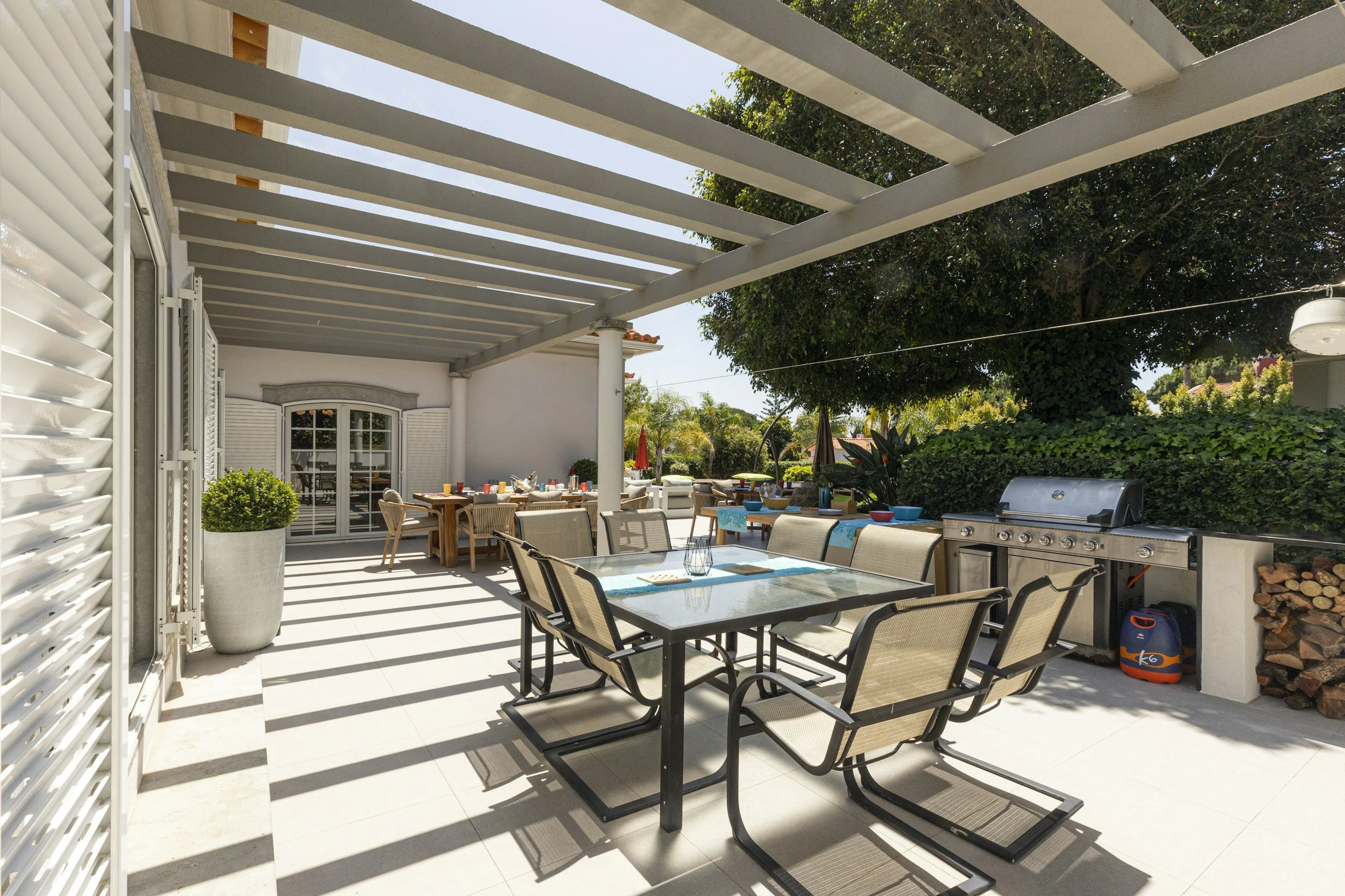 Outdoor patio with dining table, chairs, a barbecue grill, potted plant, and a white pergola with shadows, surrounded by green trees and shrubs.