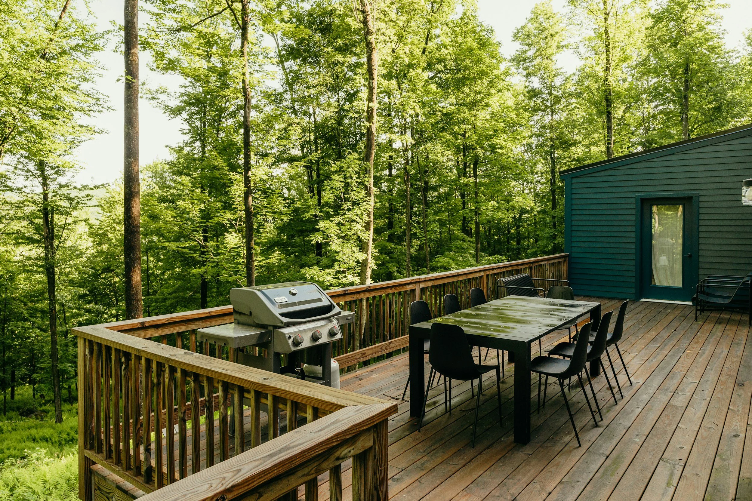 A wooden deck overlooking a lush green forest, with a black outdoor dining table and chairs, a grill, and a small blue house with a door and window on the right side.