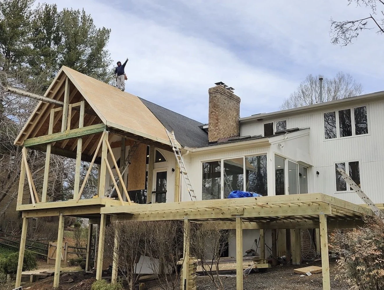 Construction of a new wooden deck and roof extension on a white house, with a worker standing on the roof and a ladder leaning against the house.