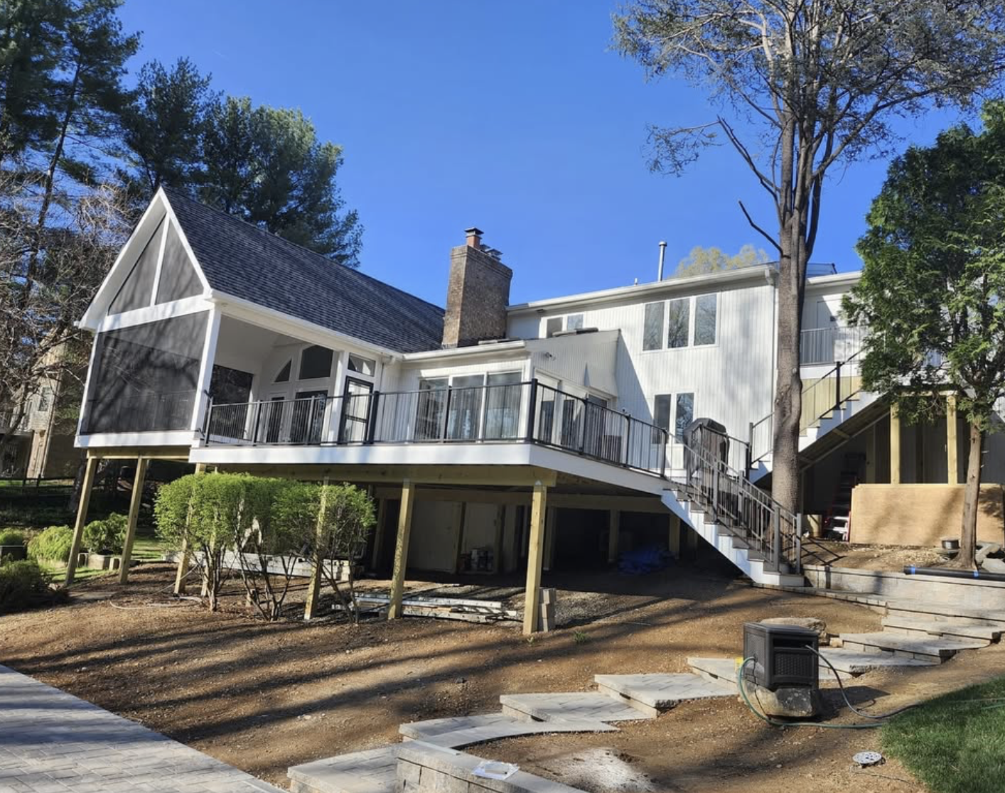 A two-story house with an upper deck, surrounded by trees, with stairs leading up to the deck. The house has a mix of siding and a steep roof, with a chimney. The yard appears to be under landscaping or renovation, with soil, some bushes, and construction materials visible.