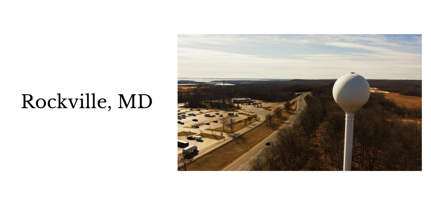A water tower on the edge of a highway near a parking lot in Rockville, Maryland, with a rural landscape in the background.