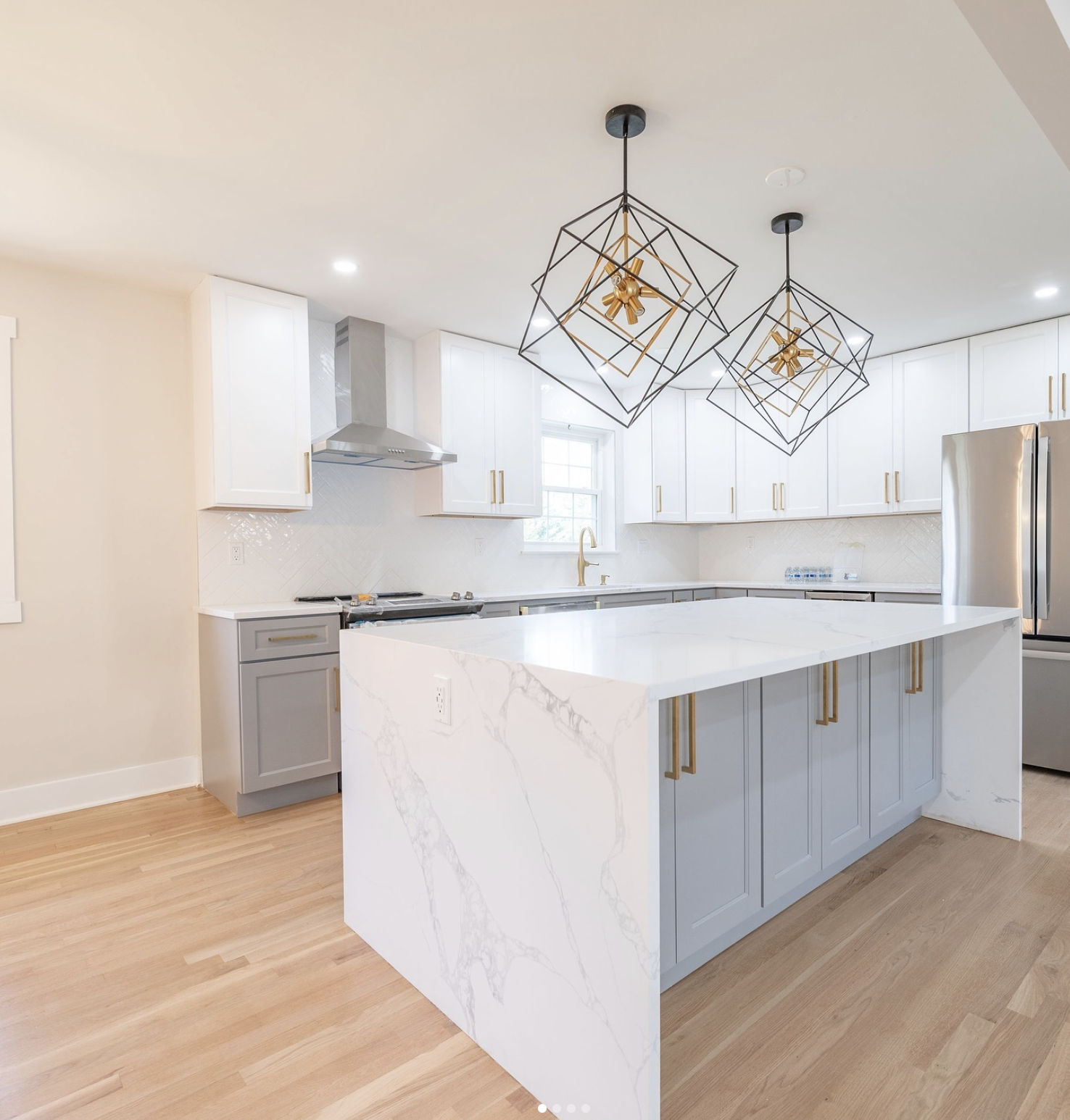 Modern kitchen with white and gray cabinets, a large white marble island, gold handles, a gold faucet, pendant lights, a stainless steel refrigerator, and a window above the sink.