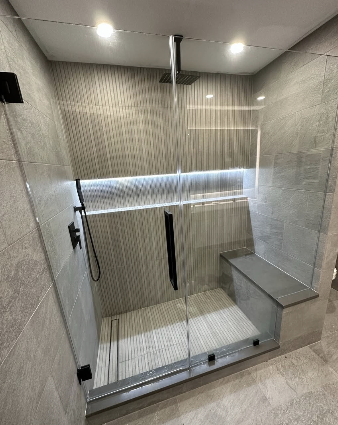 Modern walk-in shower with glass enclosure, rain shower head, and bench, with tiled walls and ceiling lights.