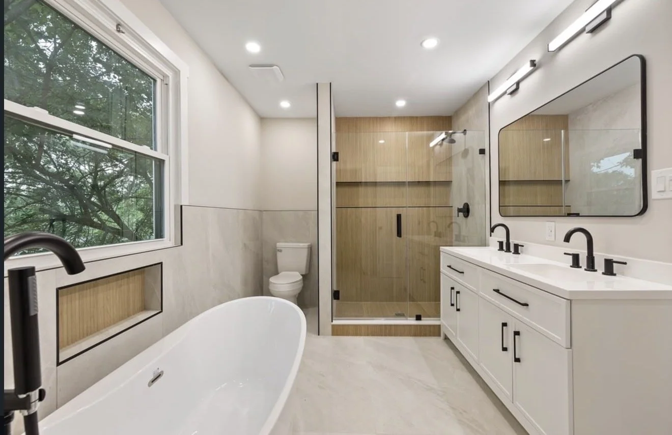 Modern bathroom with a large window, white freestanding bathtub, double vanity with black fixtures, mirror, and a glass-enclosed shower with wooden accents.
