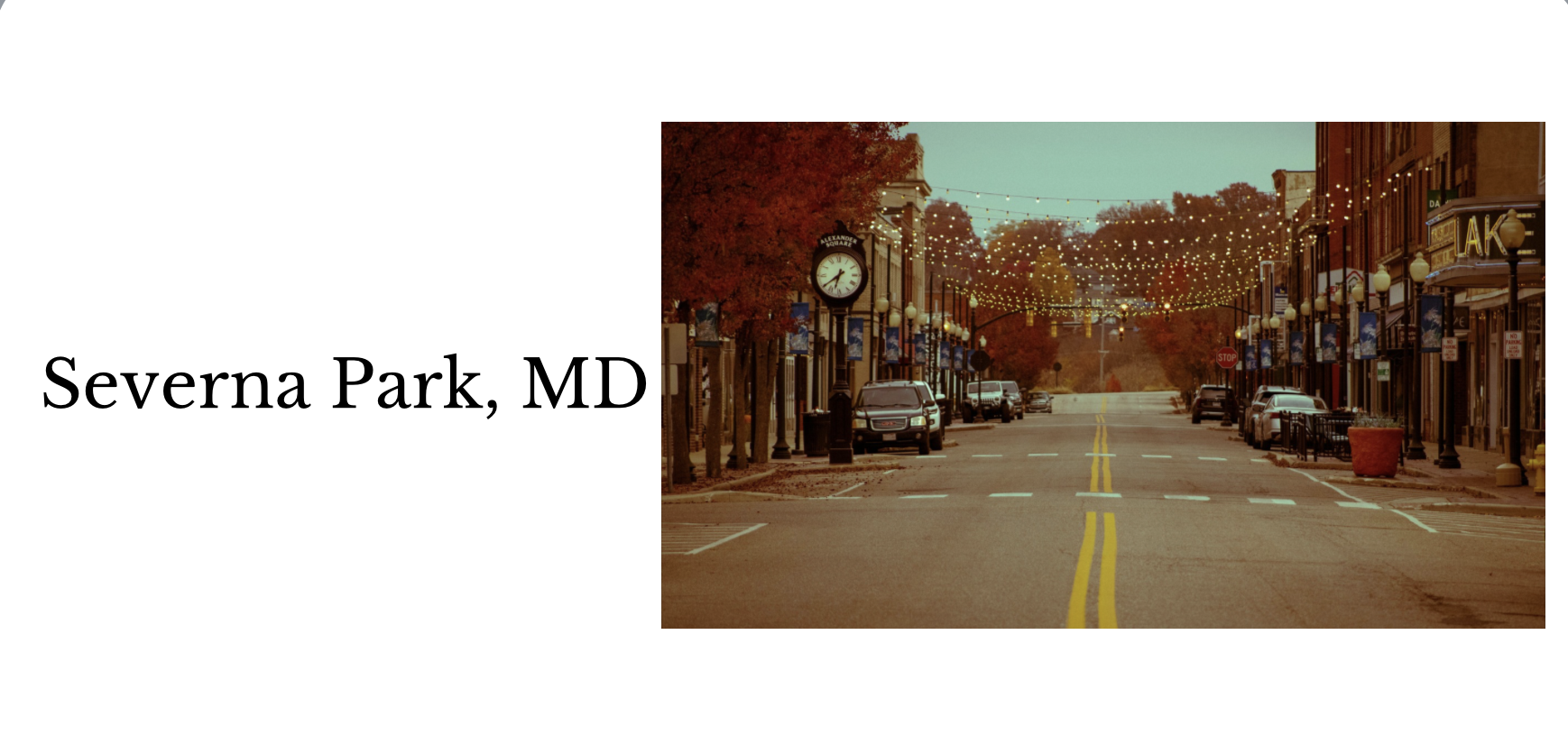 Empty street in Severna Park, MD, with parked cars, trees with autumn foliage, string lights overhead, and storefronts on both sides.