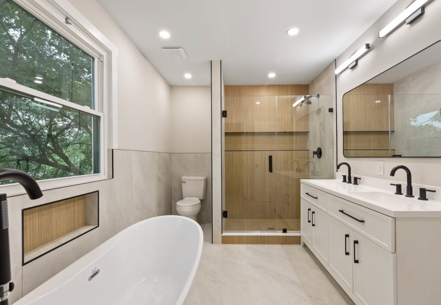 Modern bathroom with a white bathtub, large window, toilet, glass-enclosed shower, double vanity with black fixtures, large mirror, wooden cabinets, and neutral tile flooring.