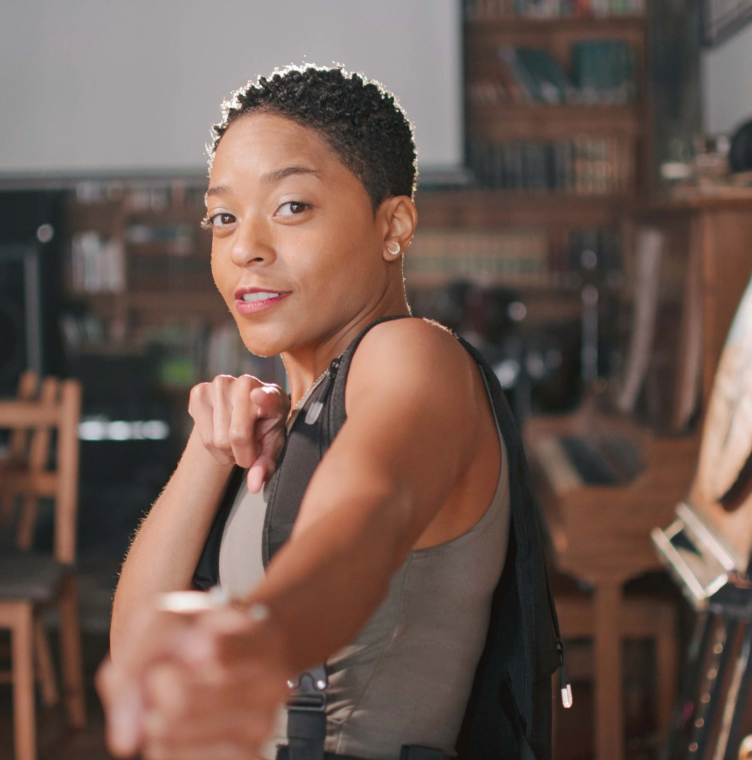 Close-up of a woman with short curly hair, wearing a black top and earrings, smiling and pointing towards the camera in an indoor setting with wooden furniture in the background.