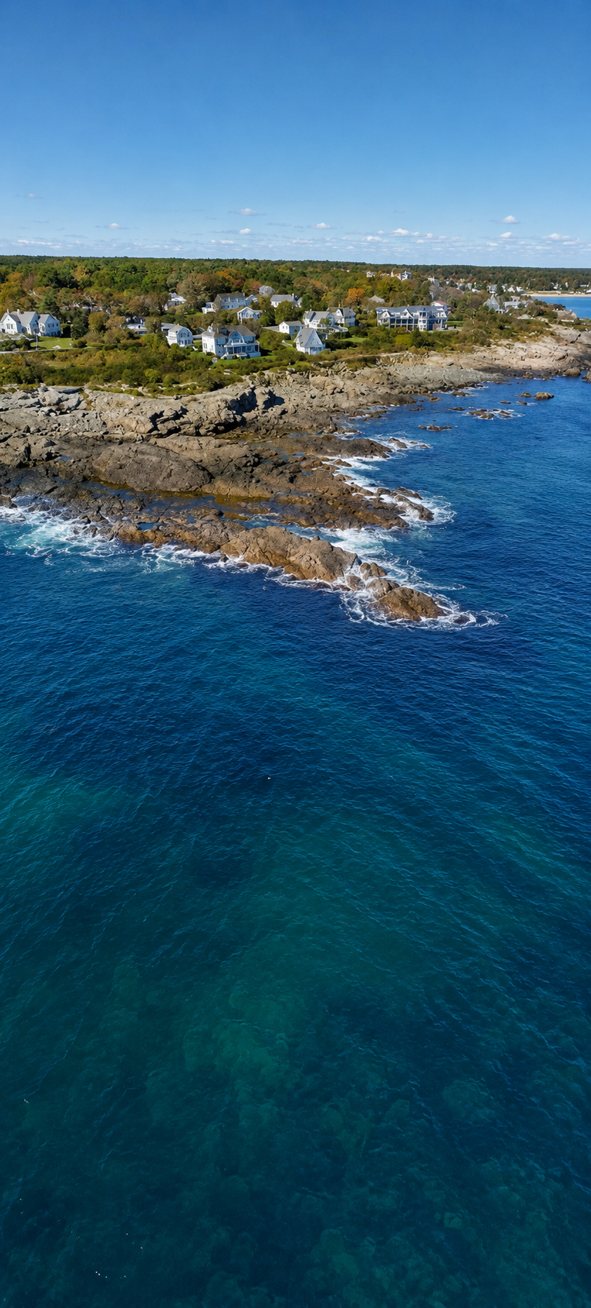 Aerial view of a coastal neighborhood with white houses and rocky shoreline, blue ocean water, and a clear sky.
