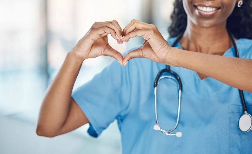 A healthcare professional in scrubs with a stethoscope making a heart shape with her hands.