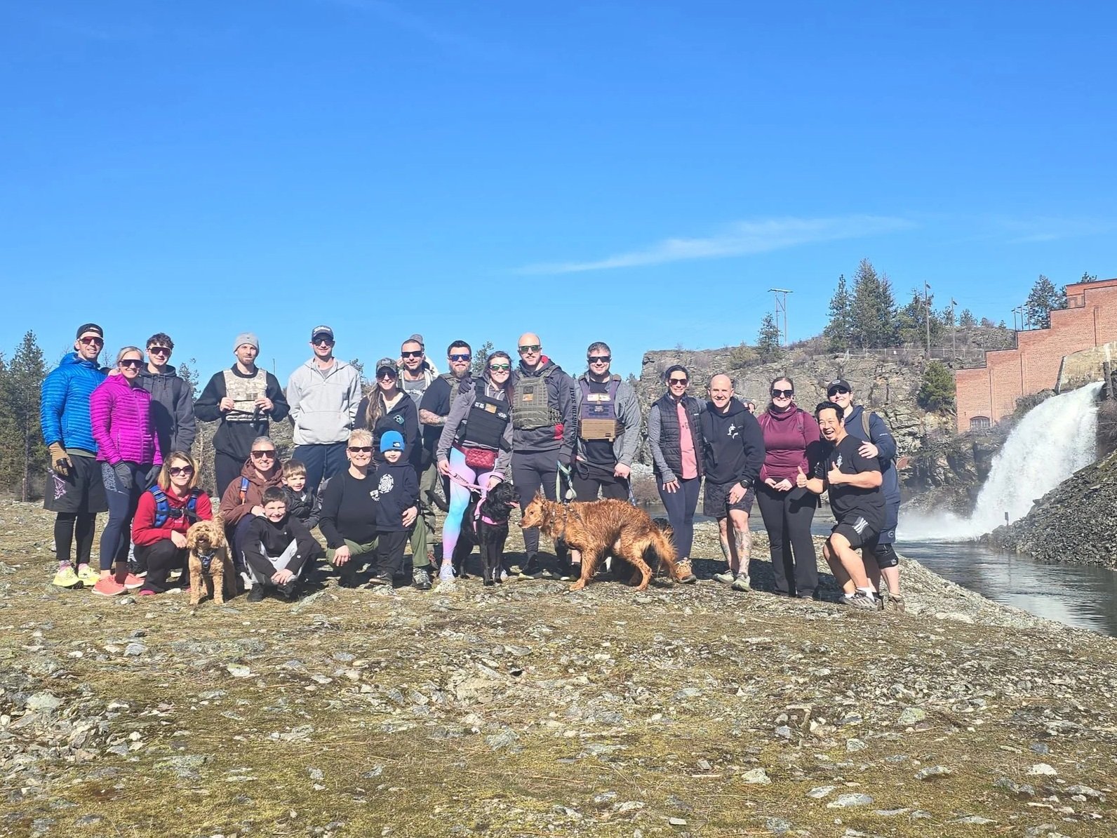 Large group of people, including kids and dogs, posing outdoors near a waterfall and rocky terrain under a clear blue sky.