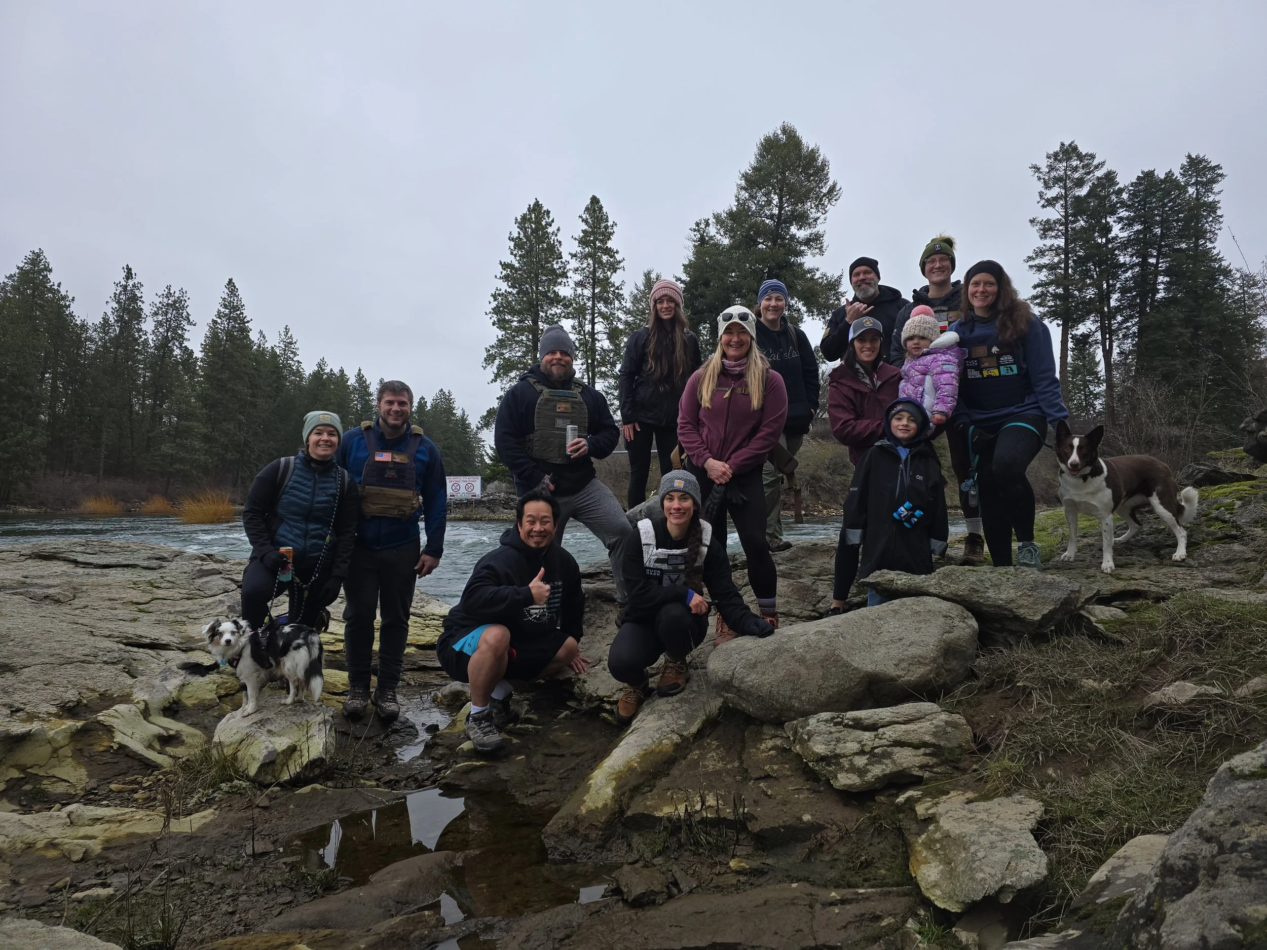 A group of people standing and kneeling on large rocks near a river in a wooded area, with trees in the background, during overcast weather. Some pets are also present, including a black and white dog in the foreground.