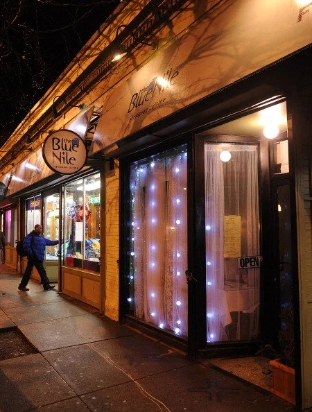 Nighttime exterior of a storefront with a lit sign reading 'the Blue Nile,' decorated with string lights, and an open door with a yellow notice inside.