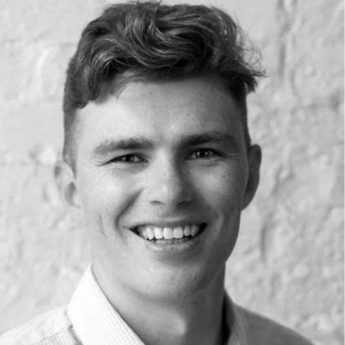 A young man with short wavy hair smiling, wearing a collared shirt, standing against a textured brick wall.