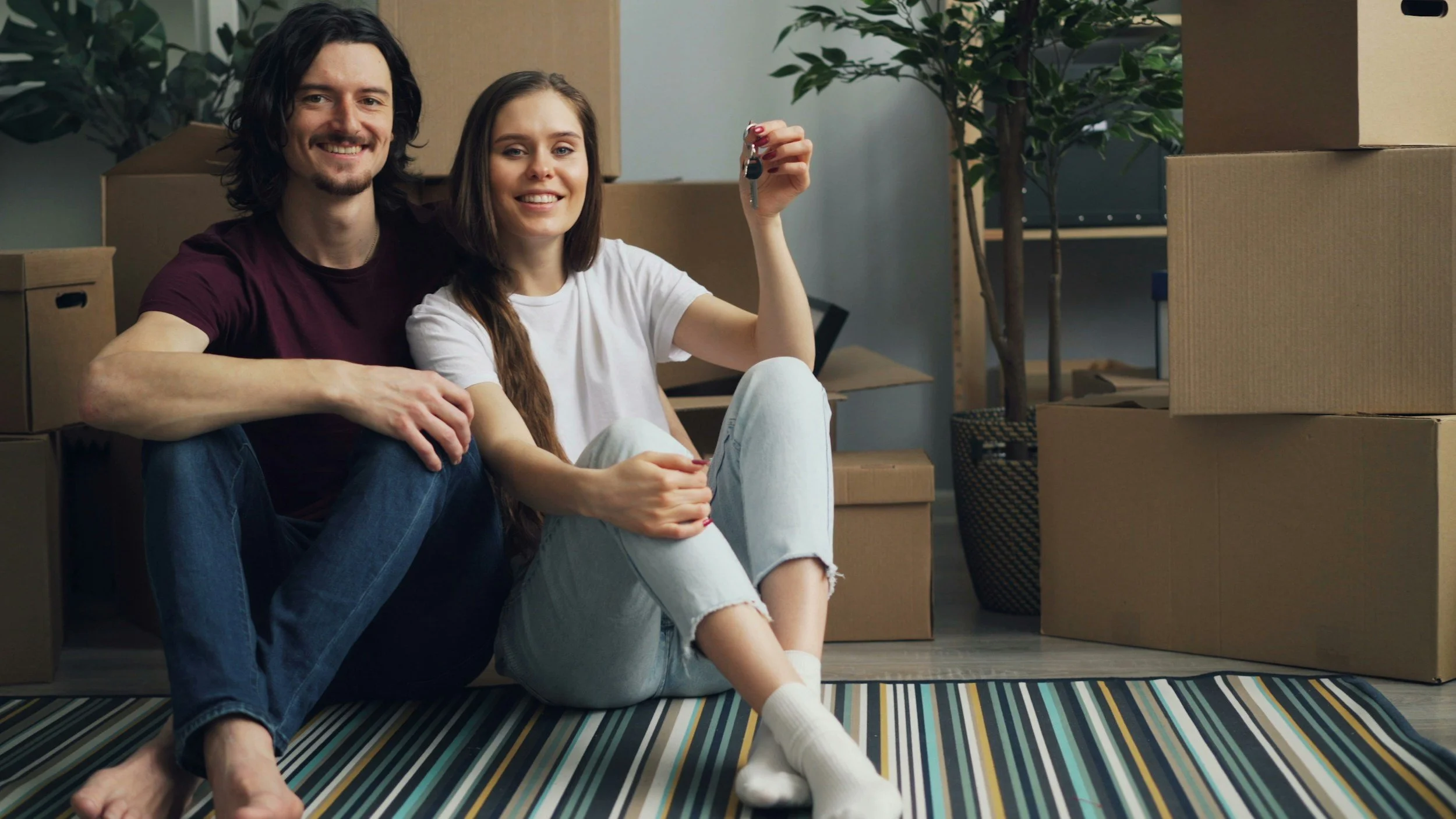 A smiling young man with long dark hair and a beard, wearing a maroon t-shirt and jeans, sitting beside a young woman with long brown hair, wearing a white t-shirt and light-colored jeans, holding a set of keys. They are surrounded by moving boxes in a room with potted plants and a striped rug.