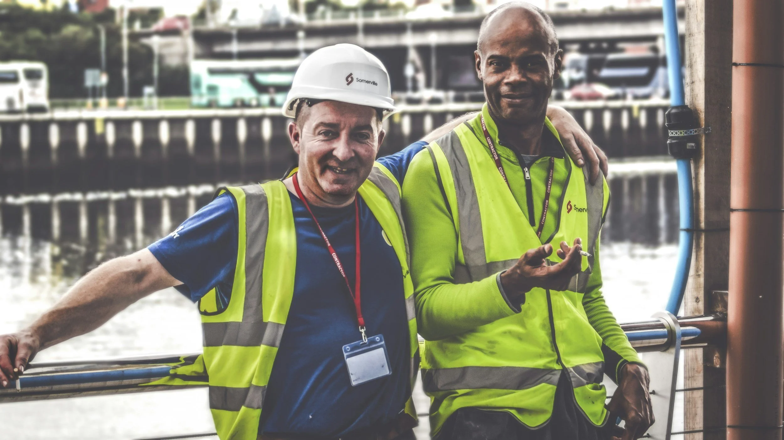 Two men, one wearing a white hard hat and both in neon yellow safety vests, are standing outdoors near a body of water with boats and a bridge in the background, smiling at the camera.
