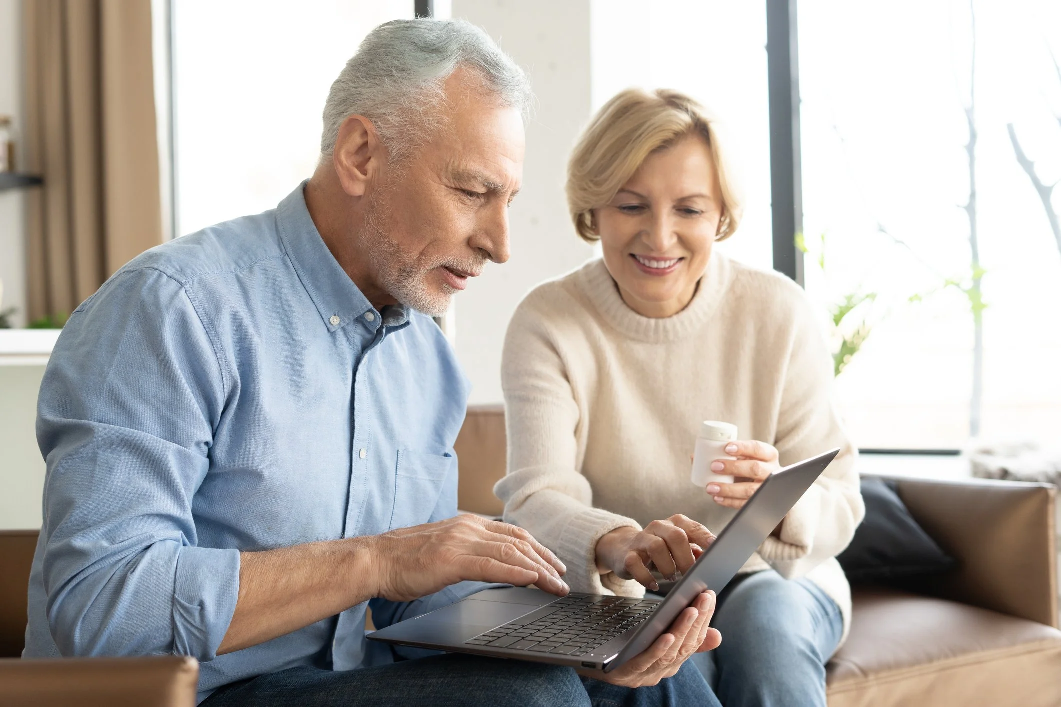An elderly man and woman sitting together on a couch, looking at a tablet device. The woman is also holding a pill bottle and a smartphone, both smiling and engaging with the devices.