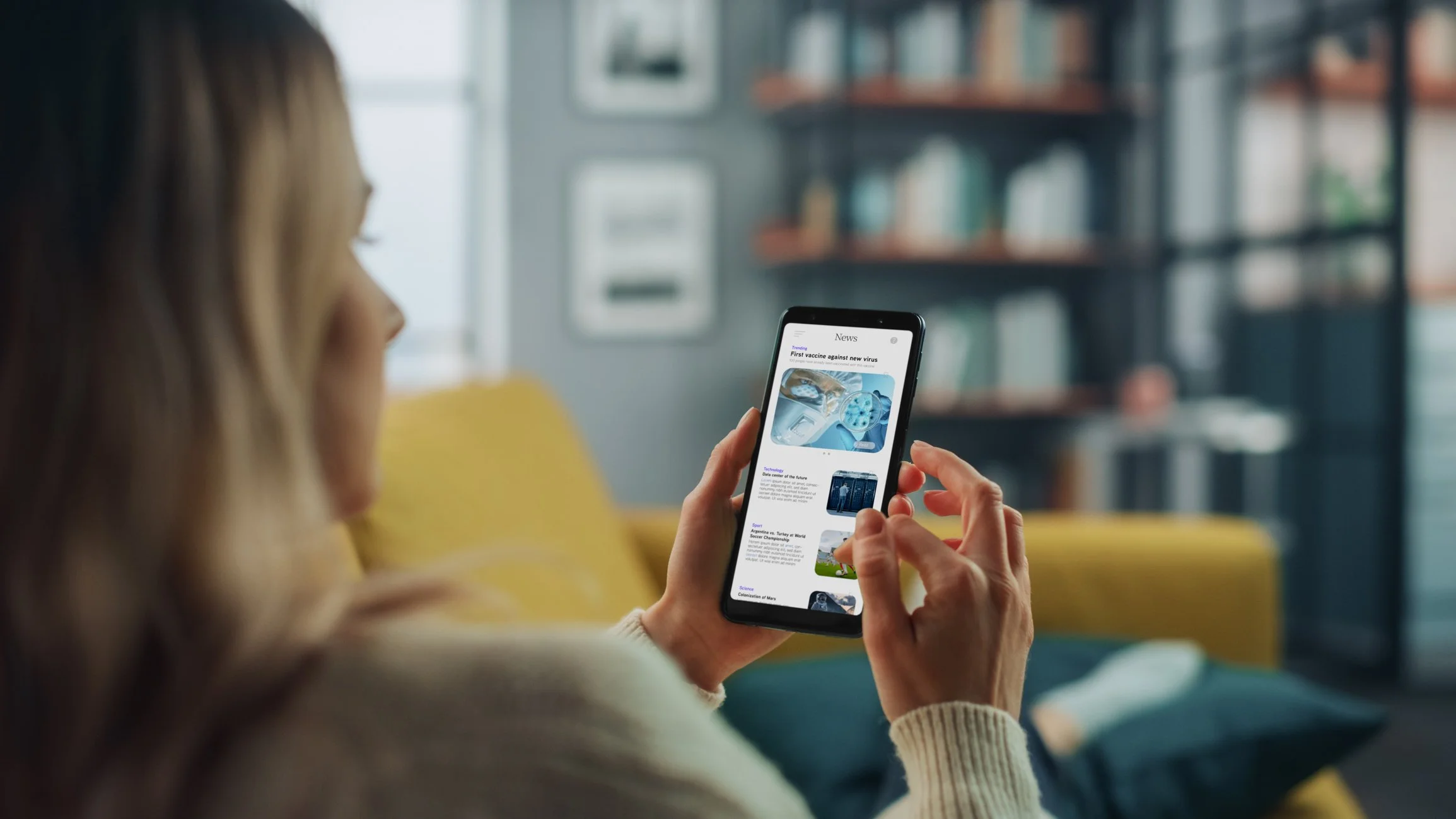 A woman with blonde hair wearing glasses sits on a yellow couch in a room with bookshelves, holding a smartphone and reading a news article.