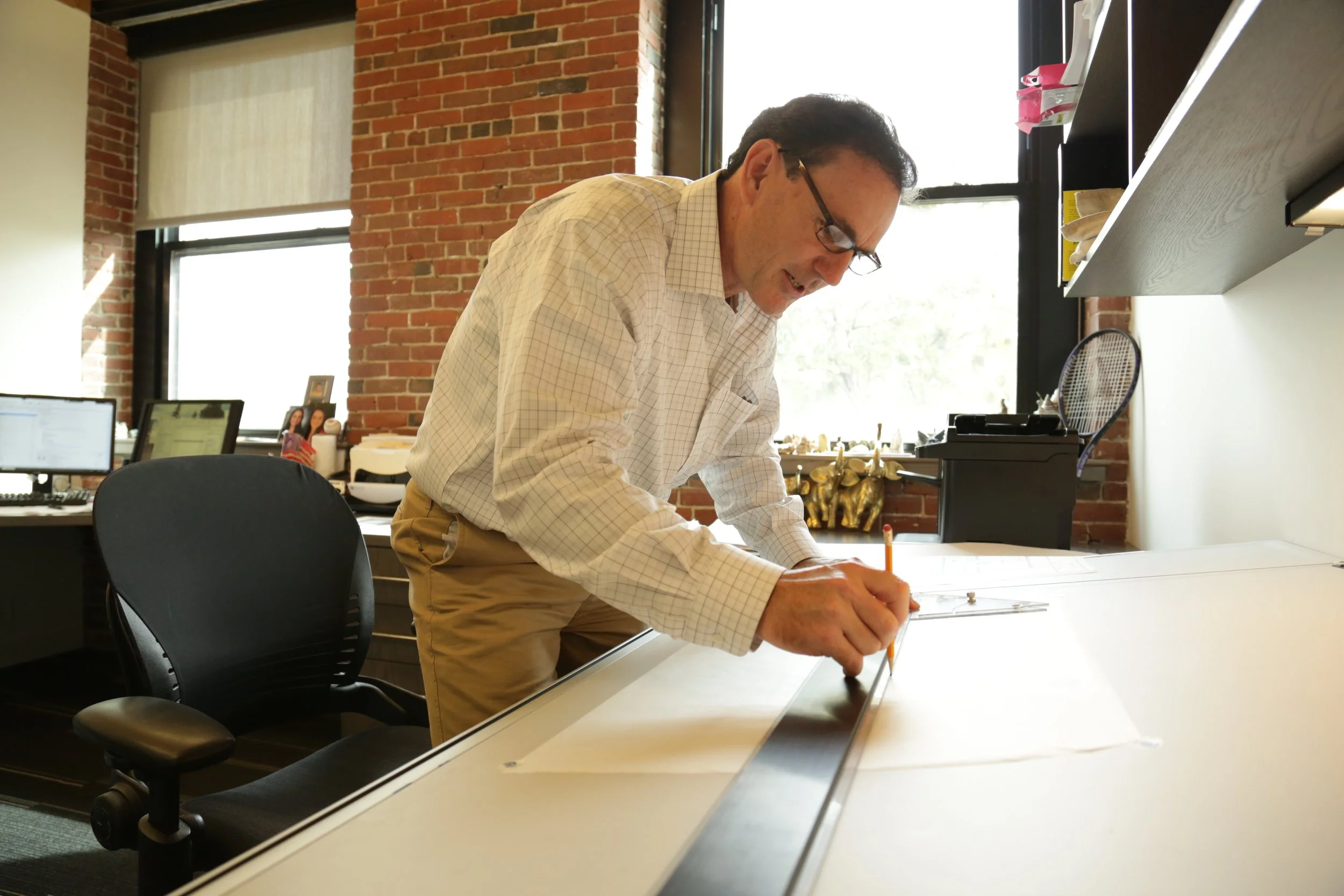 A man with dark hair and glasses wearing a white checked shirt and tan pants is leaning over a large white table, writing or drawing with a pencil. The room has exposed brick walls and large windows letting in natural light, with office desks, monitors, and framed pictures in the background.