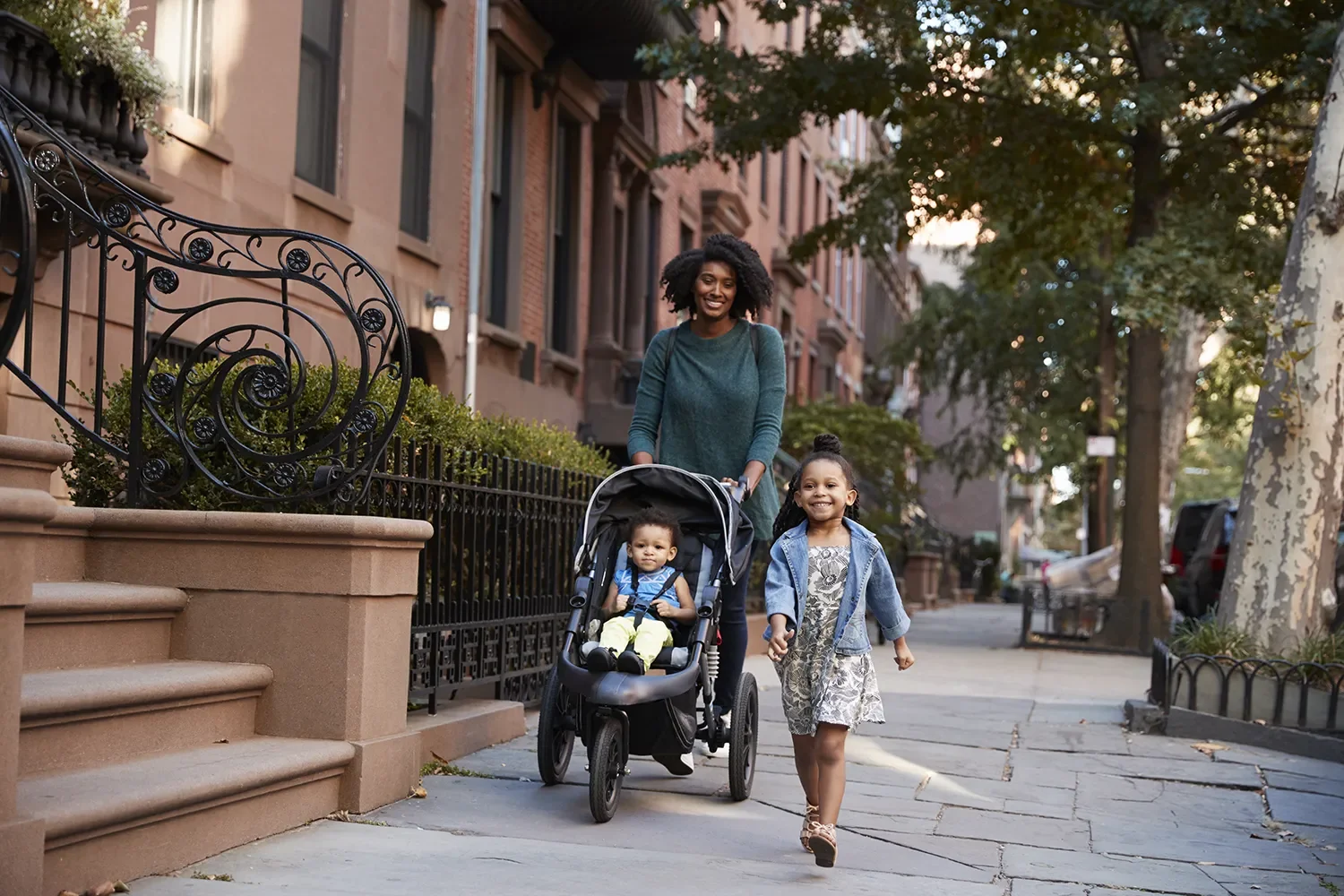 A woman walking with two young children, one in a stroller and one walking beside her, on a city sidewalk during sunset, with brownstone buildings and trees in the background.