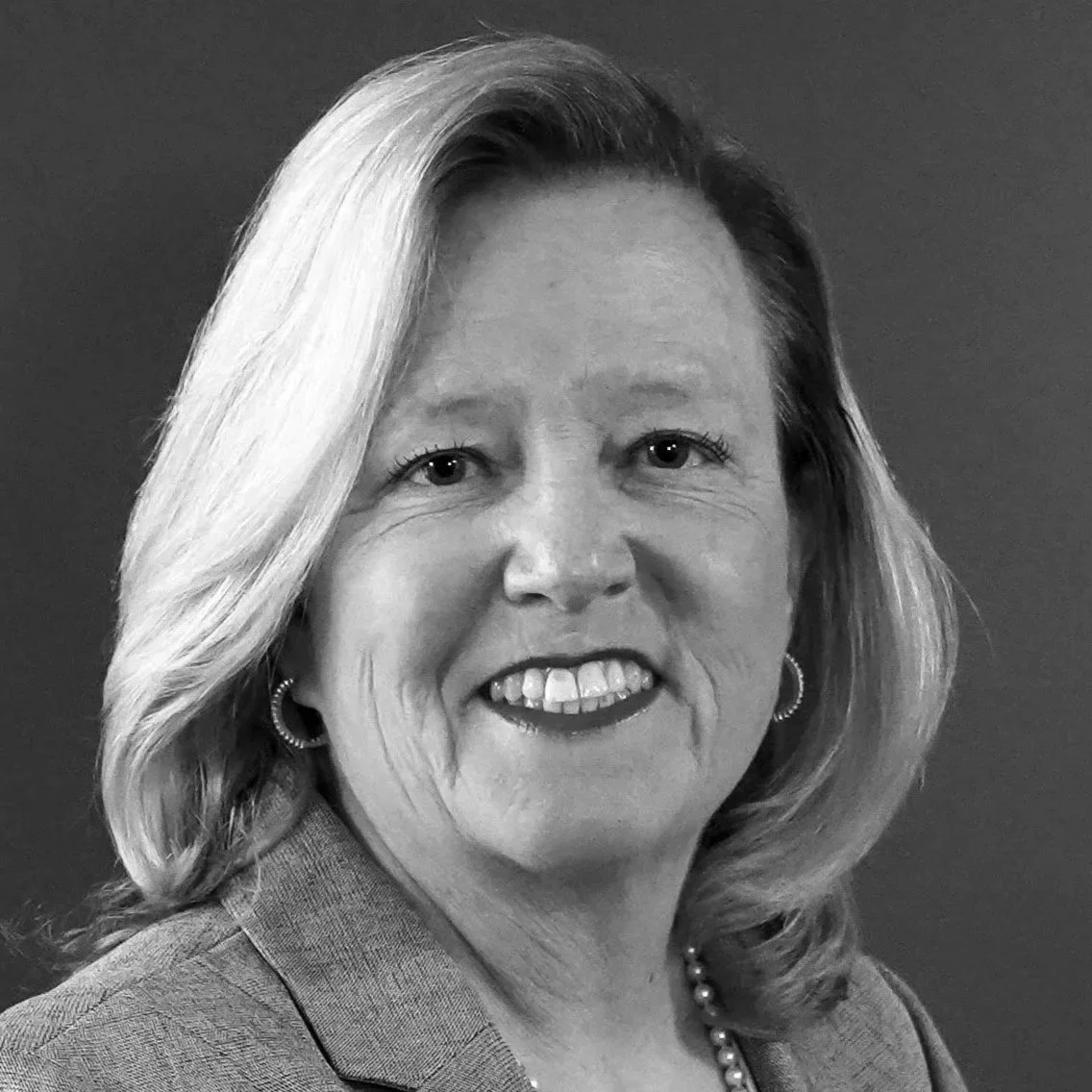 Black and white photo of a woman with shoulder-length blond hair, wearing a blazer, earrings, and a pearl necklace, smiling at the camera.
