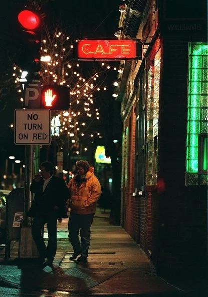 Nighttime city street scene with pedestrians, streetlights, and a red illuminated 'CAFE' sign.