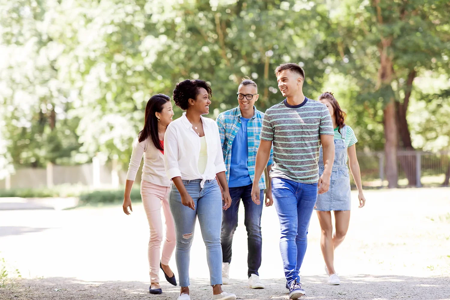 A diverse group of five young adults walking together outdoors in a park, smiling and engaging in conversation.