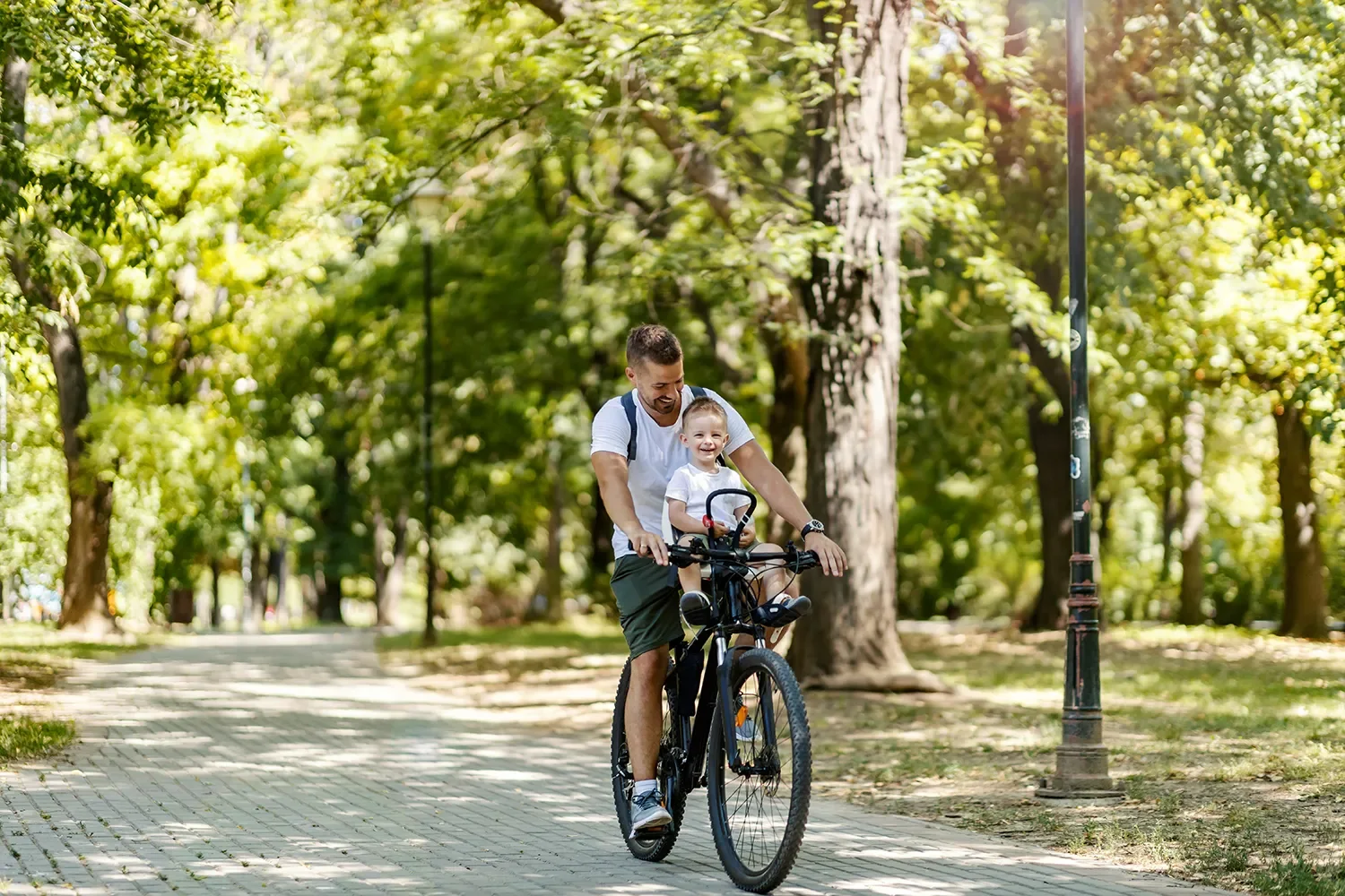 A father and young child riding a bicycle together in a park with green trees and a pathway