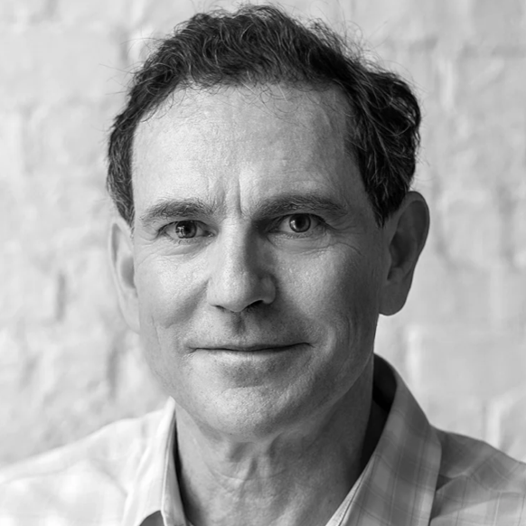 Black and white portrait of a middle-aged man with short dark hair, wearing a checkered collared shirt, standing against a light-colored brick wall.