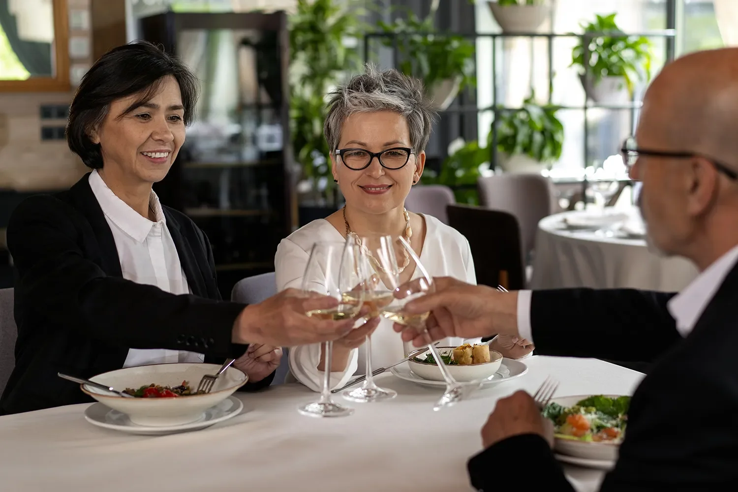 Three middle-aged adults, two women and one man, dining together at a restaurant, toasting with white wine glasses. They are seated at a table with plates of food, smiling and engaging in celebration.