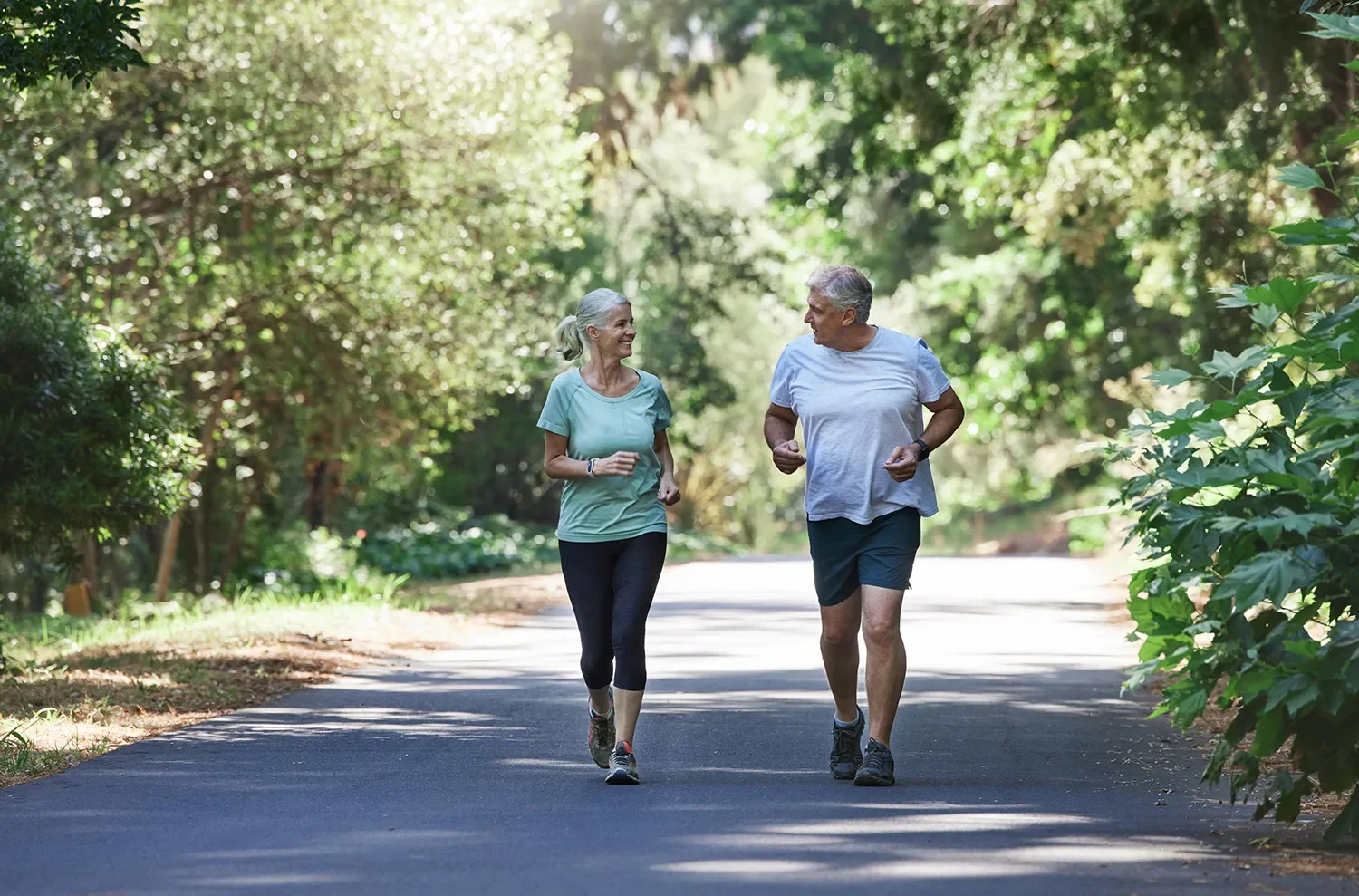 Older man and woman jogging together on a shaded path in a wooded area, smiling and talking.