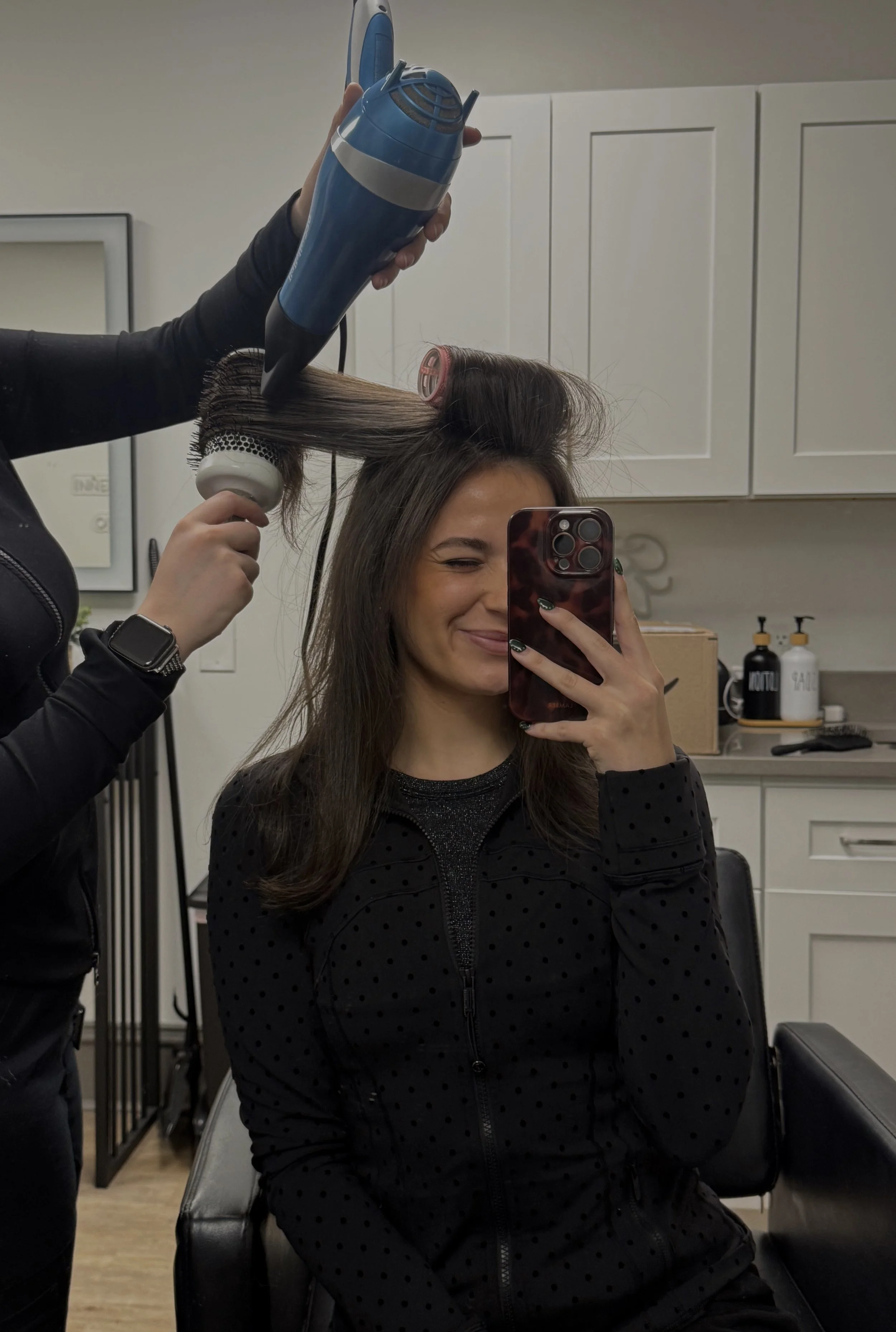 A woman with long brown hair is sitting on a black chair and taking a selfie in a mirror. A hairstylist is blow-drying her hair with a blow dryer and a round brush. The woman is smiling and winking. The setting appears to be a salon with white cabinets and bottles in the background.