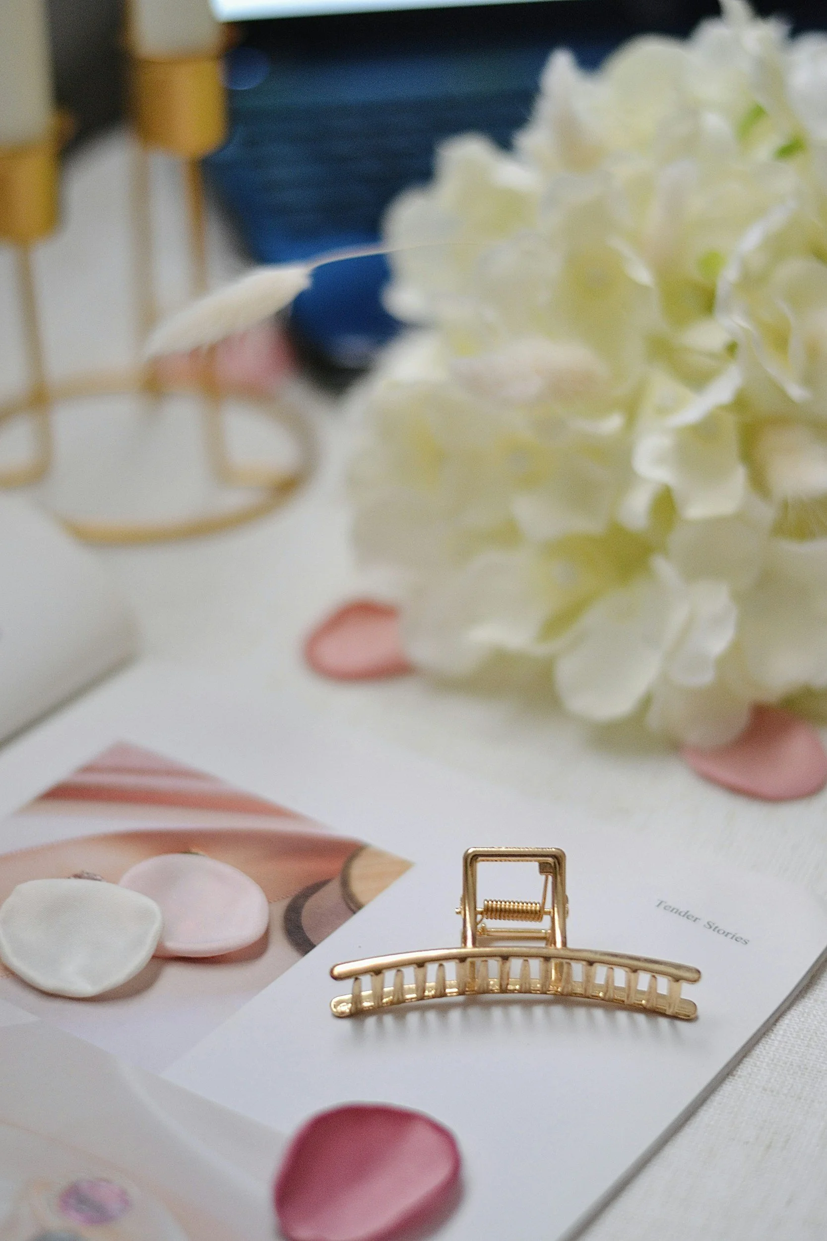 Close-up of a display with a gold decorative hair clip, a white hydrangea flower, pink and white petals, and a magazine with images of makeup products and the word 'Tender Stories' visible.