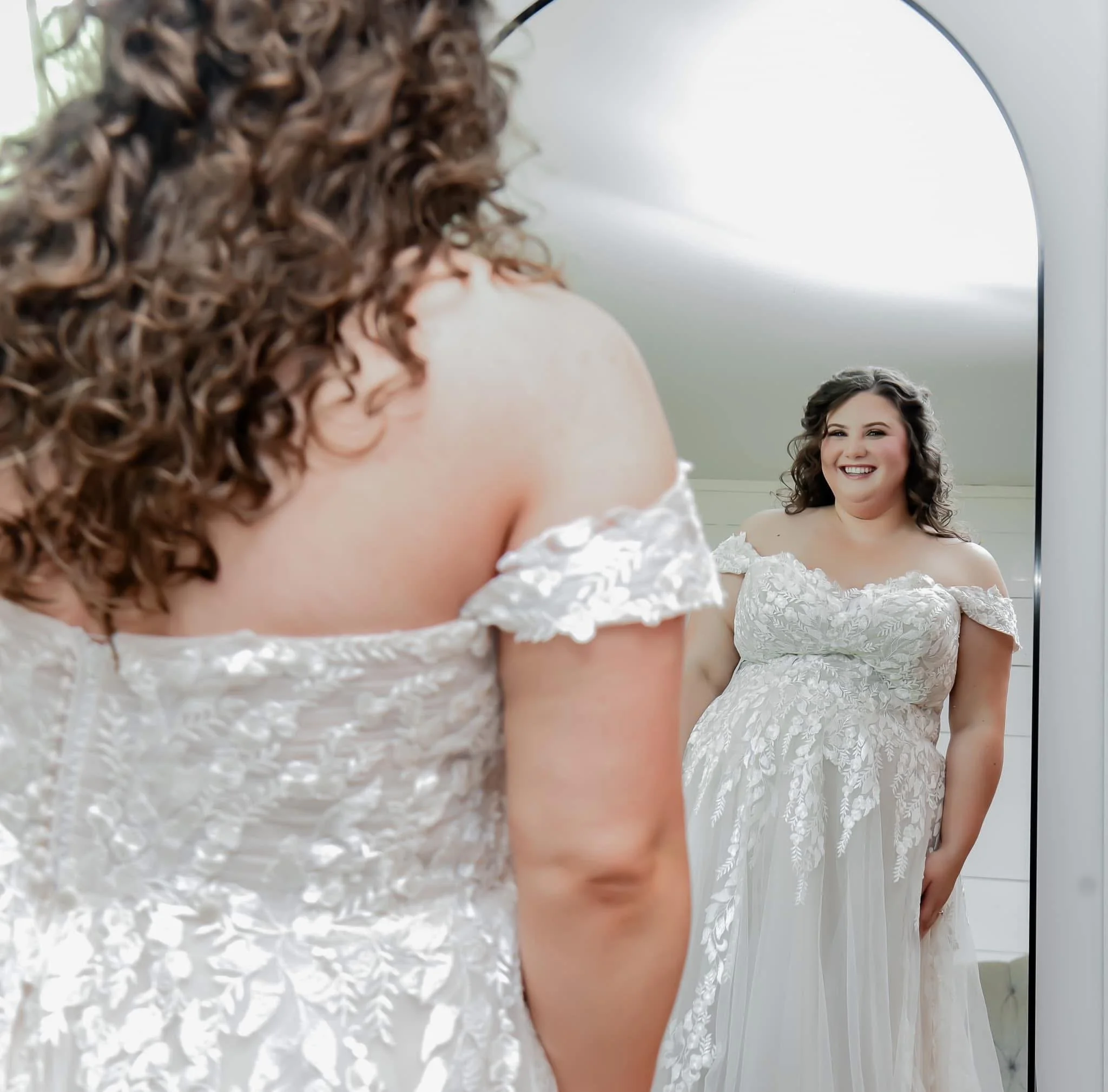 A woman with curly hair smiling in front of a mirror, wearing a white off-shoulder dress, in a bridal or formal setting.