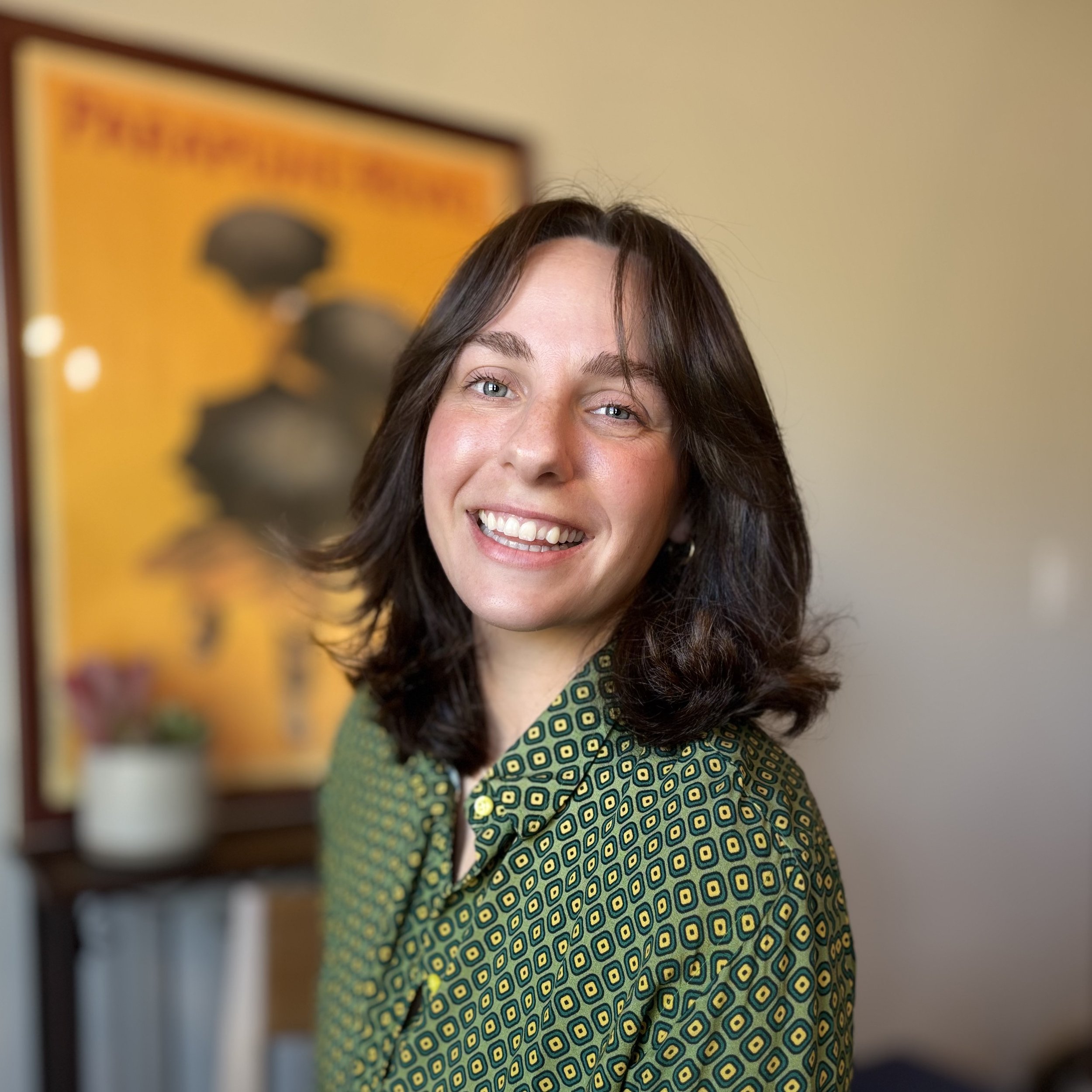 A woman with blue eyes and dark wavy hair smiling, wearing a green patterned button down shirt, standing in front of a yellow framed art piece with black circles and orange background.