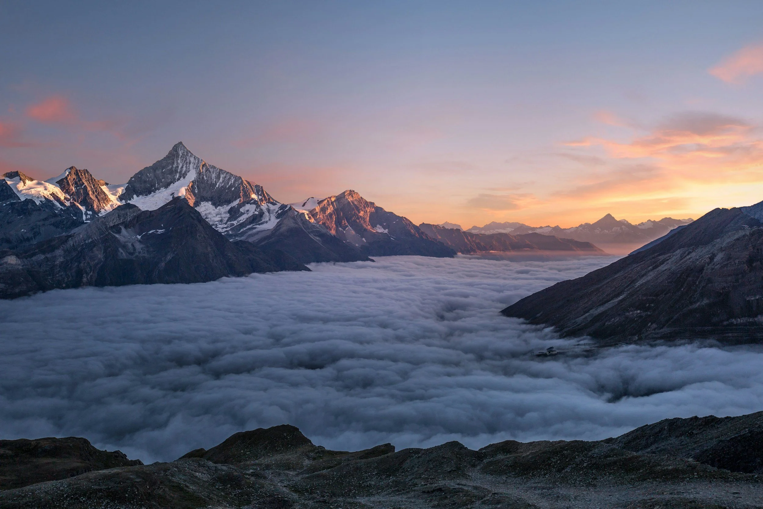 Snow-capped mountains at sunrise with a layer of clouds below and the sky showing soft pastel colors.