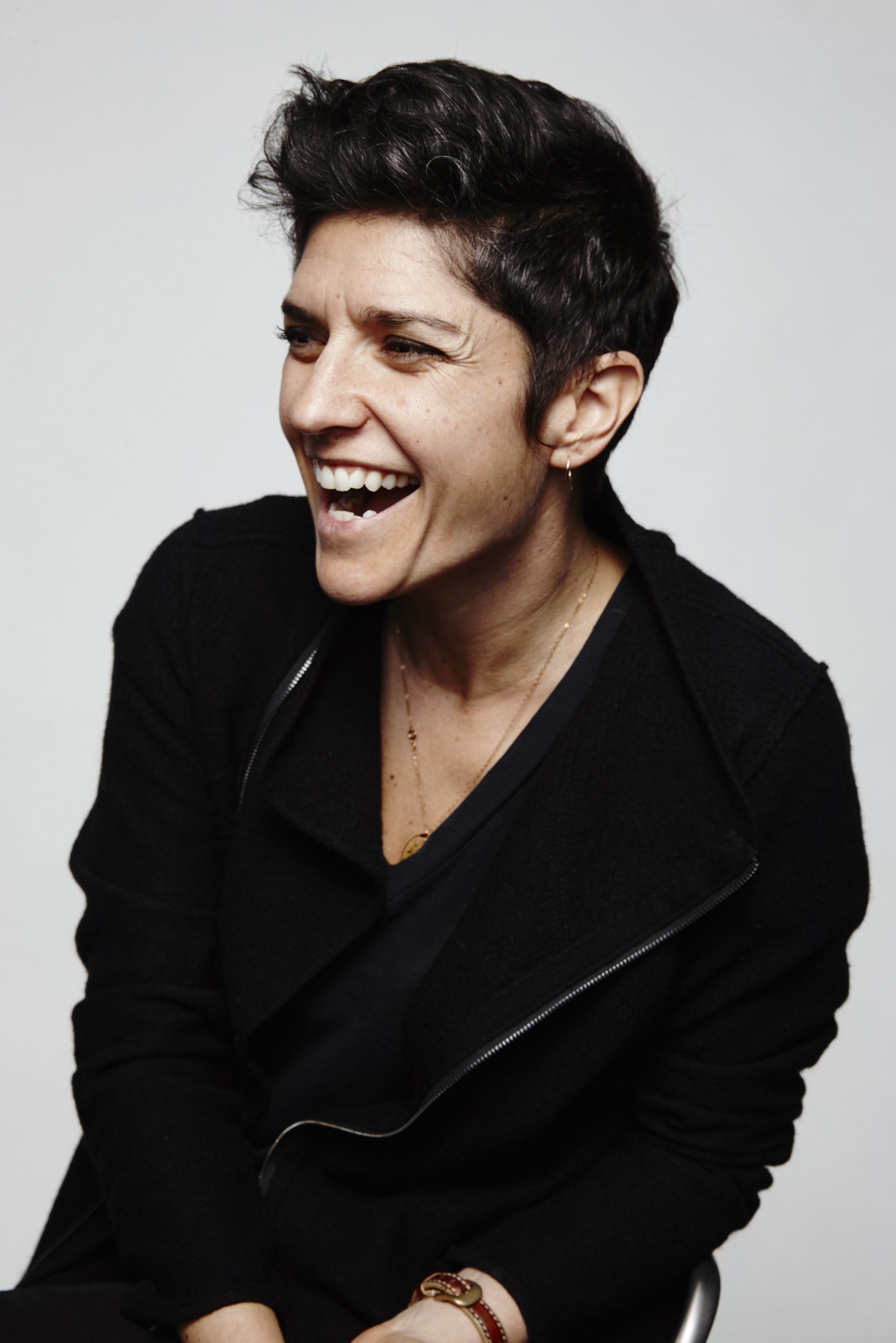 A woman with short dark hair and light skin smiling and laughing, sitting against a white background. She is wearing a black top, gold jewelry, and a brown bracelet.