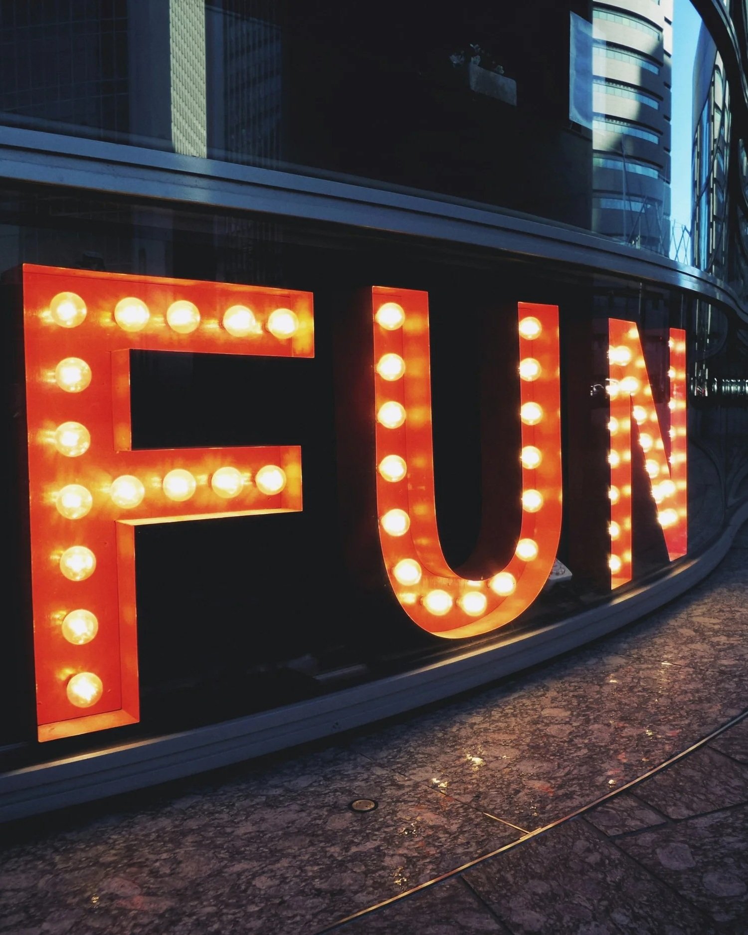 Close-up of illuminated retro-style sign spelling 'FUN' with round light bulbs, mounted on a glossy dark background, with reflections of modern buildings and a street surface below.