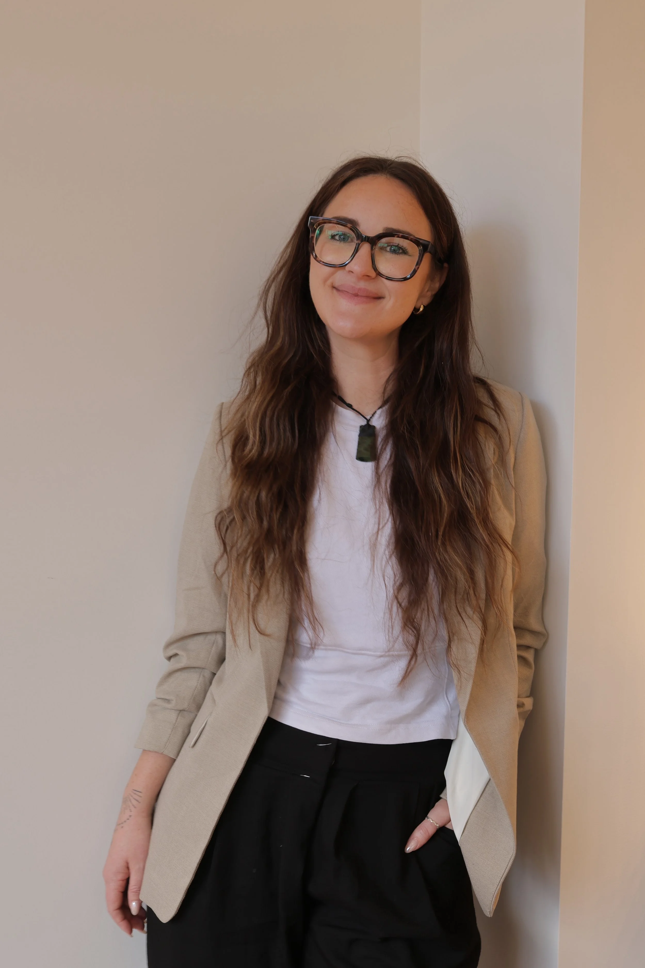 A woman psychologist with long wavy brown hair wearing a beige blazer, white t-shirt, and black pants, standing against a neutral-colored wall. Wearing a pounamu and glasses, smiling.