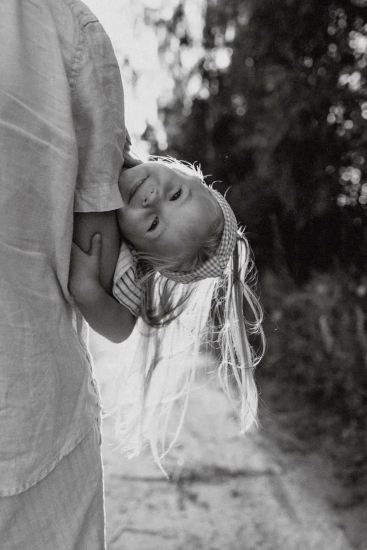 A young girl with long hair and a headband resting her head on an adult's shoulder outdoors, black-and-white photo with trees in the background.