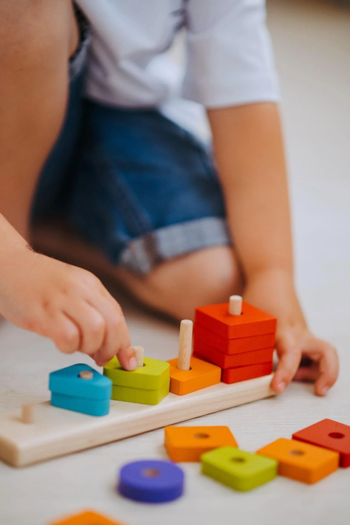 Close-up of a child playing with colorful wooden stacking toys on a white surface. The child is wearing a white shirt and dark blue shorts.