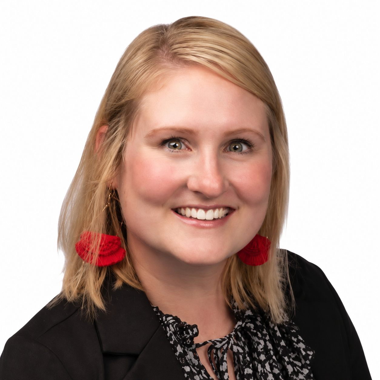 Smiling woman with blonde hair wearing red earrings, a black blazer, and a patterned blouse against a white background.